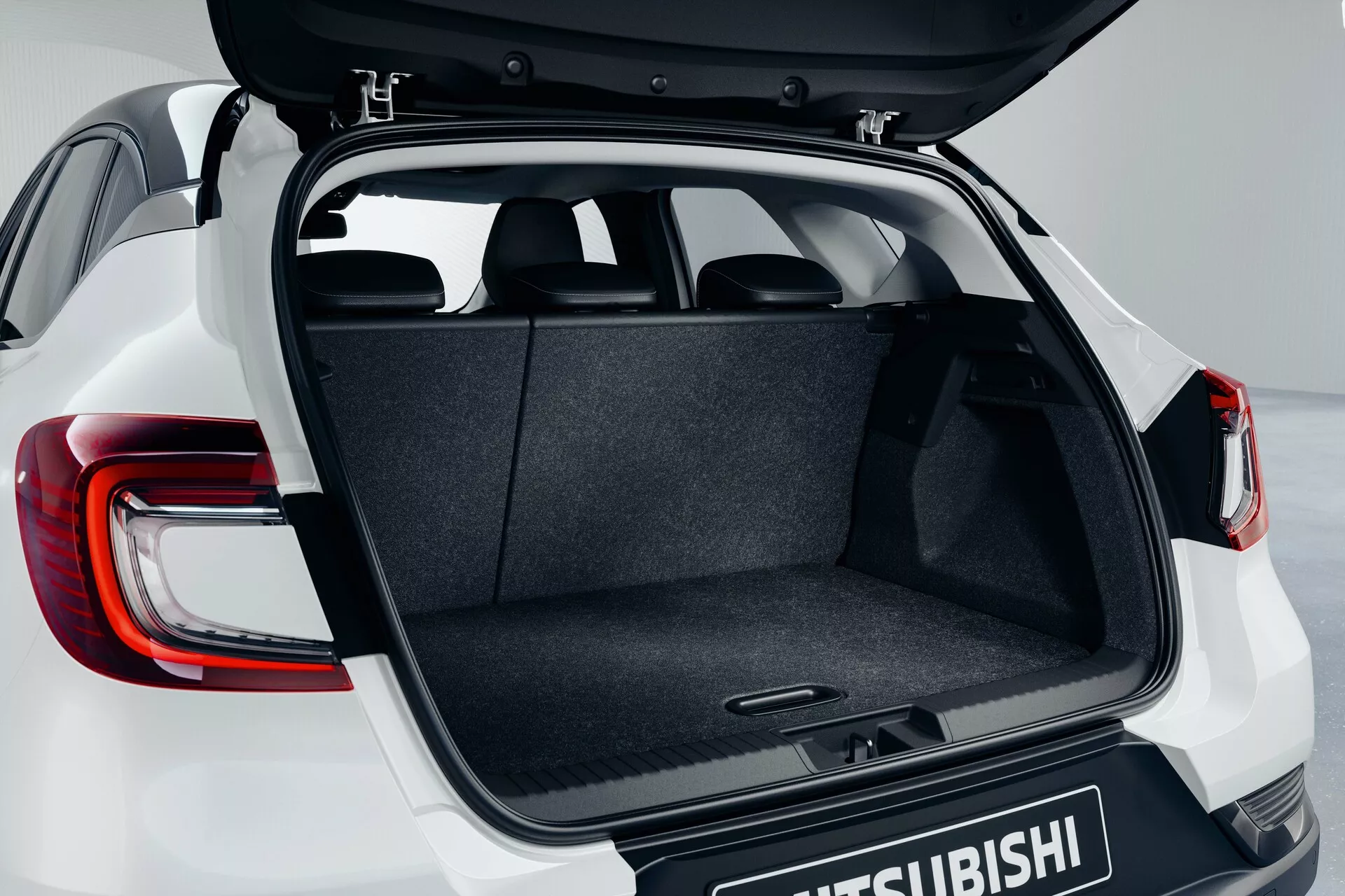 Vano posteriore del Mitsubishi ASX 2023 (2ª generazione) che mostra il portellone aperto da un'angolazione leggermente elevata, evidenziando lo spazio di carico rivestito in moquette, il meccanismo della cerniera, il pavimento piatto e le luci posteriori a LED moderne parzialmente visibili con il logo Mitsubishi sulla porta del bagagliaio.