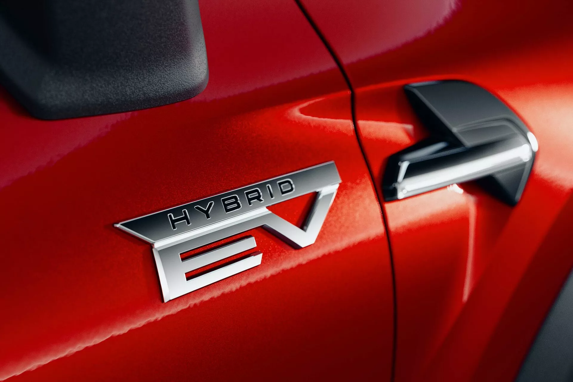 Dettaglio ravvicinato del Mitsubishi ASX 2023 (2ª generazione) in rosso, che mostra il distintivo 'HYBRID EV' che indica la tecnologia del veicolo ibrido elettrico. Il distintivo 'HYBRID EV' è centrato, mettendo in risalto la finitura metallica e le precise lettere. Il 'EV' presenta un design distintivo con una freccia che attraversa la 'V'. Lo specchietto retrovisore laterale è parzialmente visibile nell'angolo in alto a destra, suggerendo caratteristiche tecnologiche aggiuntive. La finitura lucida della vernice e la cucitura stretta tra la porta del passeggero anteriore e il parafango anteriore suggeriscono un'alta qualità costruttiva.