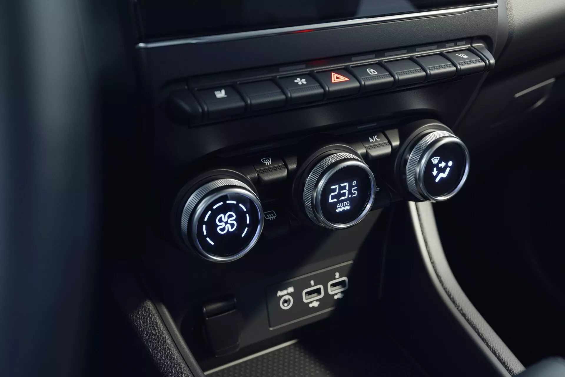 Vista ravvicinata del sistema di controllo climatico interno della Mitsubishi ASX 2023 (seconda generazione). L'immagine, scattata da un'angolazione elevata, si concentra sulla console centrale con tre manopole di controllo della temperatura: la sinistra impostata su 'LO' e la destra su '23.5'. La manopola centrale sembra controllare la velocità o la direzione della ventola. Sopra ci sono pulsanti per sbrinamento, luci di emergenza e circolazione dell'aria. Sotto ci sono moderni porti USB. Il design è minimalista con superfici nere ed eleganti.