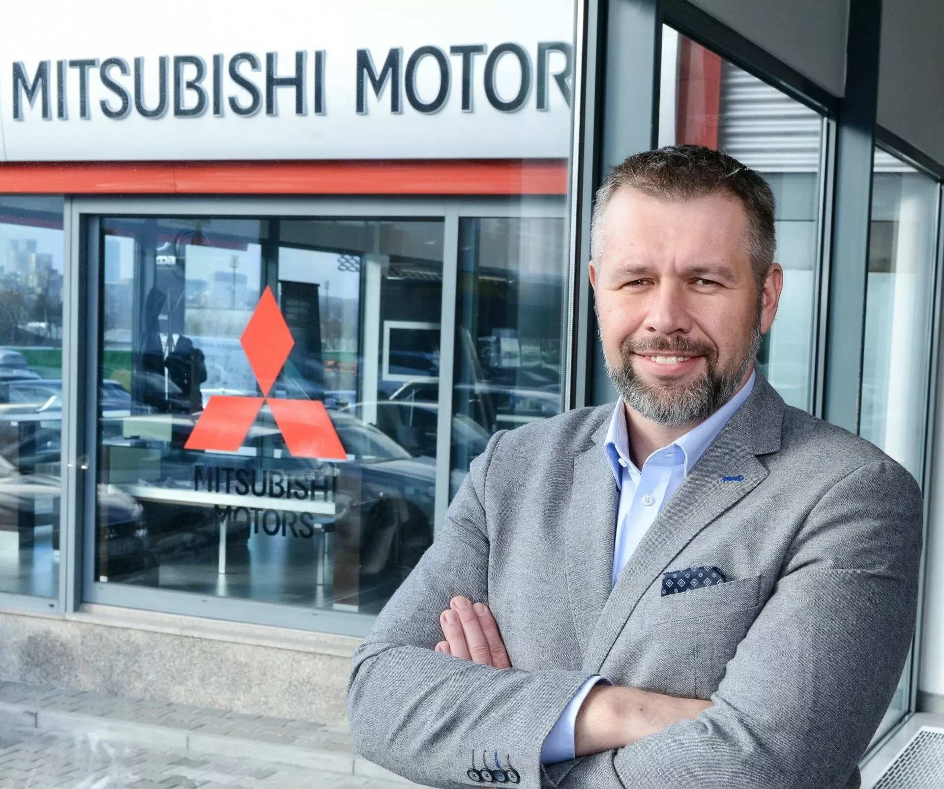 Persona in piedi con le braccia incrociate davanti a una concessionaria Mitsubishi Motors, veicolo parzialmente visibile attraverso la finestra in vetro che suggerisce un modello in esposizione sullo sfondo