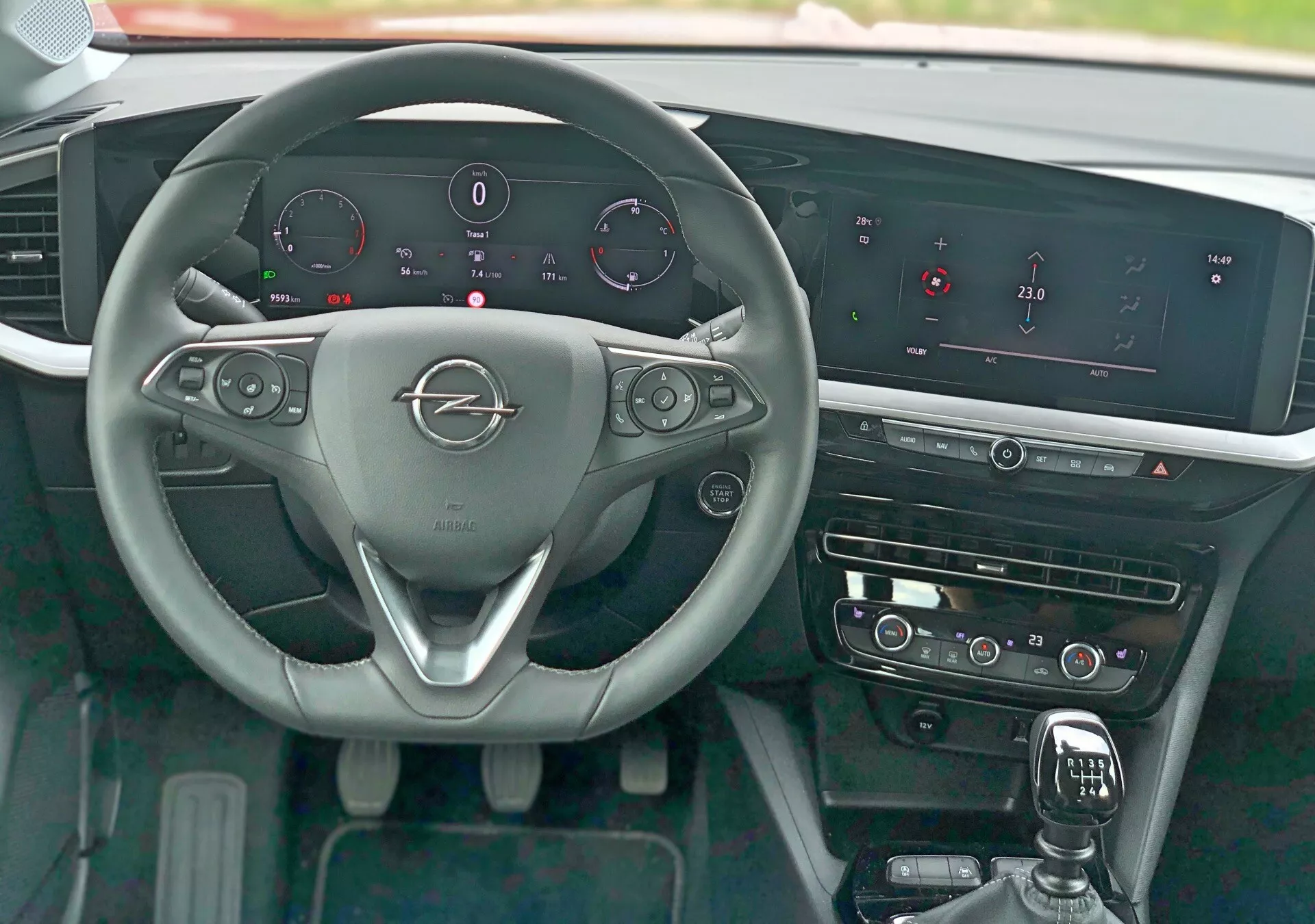 Vista dell'interno dell'opel mokka 2021 dalla prospettiva del sedile del conducente che mostra il volante a tre razze con emblema metallico del marchio, comandi montati sul volante per audio, telefono e controllo della velocità di crociera, leve del cambio, quadro strumenti digitale che visualizza velocità, giri, livello del carburante e temperatura del motore, sistema di infotainment prominente con schermo orizzontale ampio, controlli a sfioramento per HVAC, leva del cambio con copertura in pelle, elementi decorativi lucidi e pulsante di avviamento senza chiave.