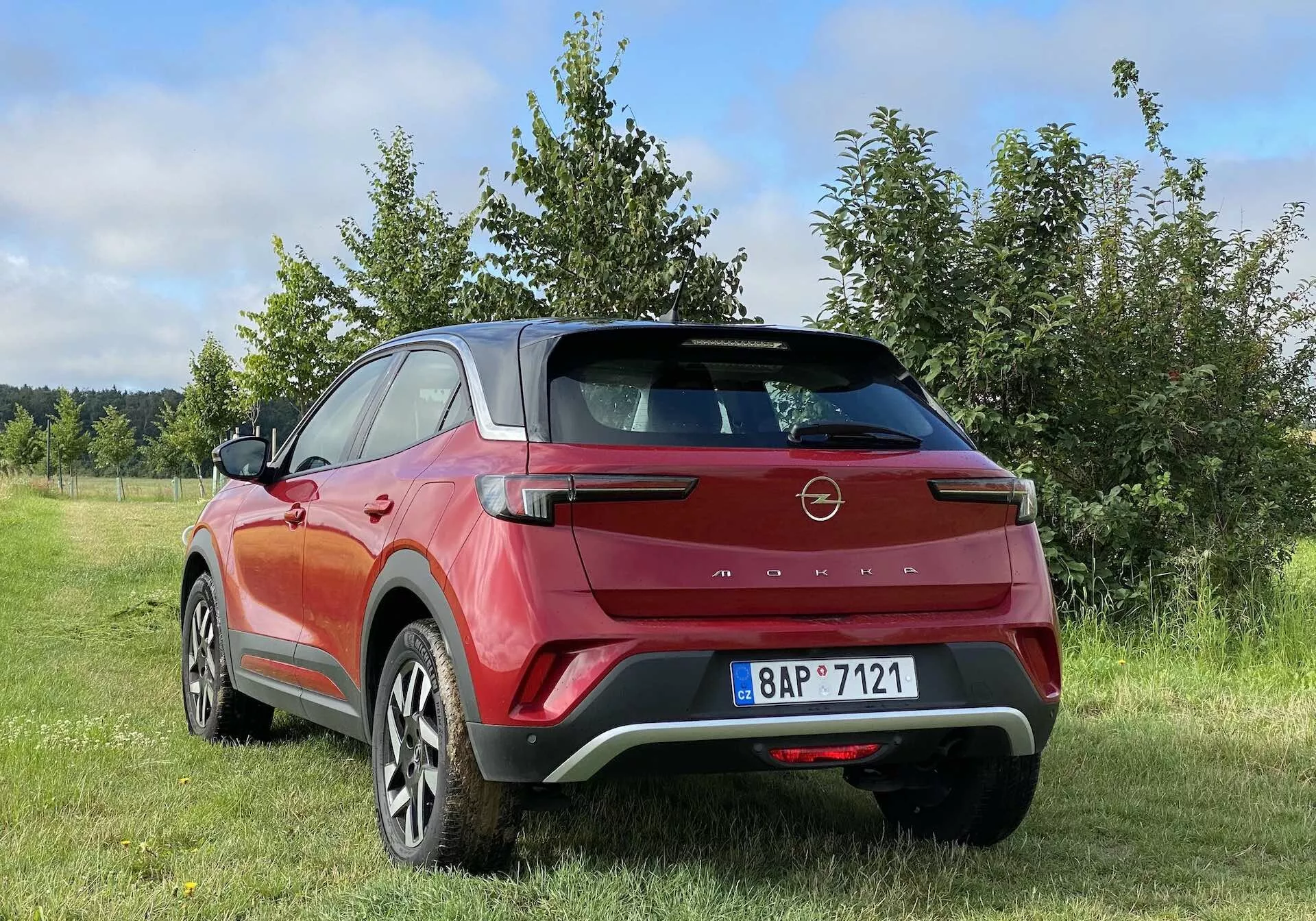 Fotografia a colori che mostra una vista posteriore tre quarti di un moderno crossover SUV Opel Mokka 2021 in rosso e nero, parcheggiato all'aperto in una zona erbosa con alberi o arbusti sullo sfondo. Gli elementi di design del veicolo includono uno spoiler posteriore aerodinamico, fanali posteriori a LED angolari, luce di frenata alta, carrozzeria bicolore, grandi cerchi in lega, maniglie delle porte a filo e rivestimenti in plastica nera. La foto è scattata da un angolo basso, mostrando l'altezza e la distanza da terra del SUV, con un cielo parzialmente nuvoloso che indica condizioni climatiche variabili.