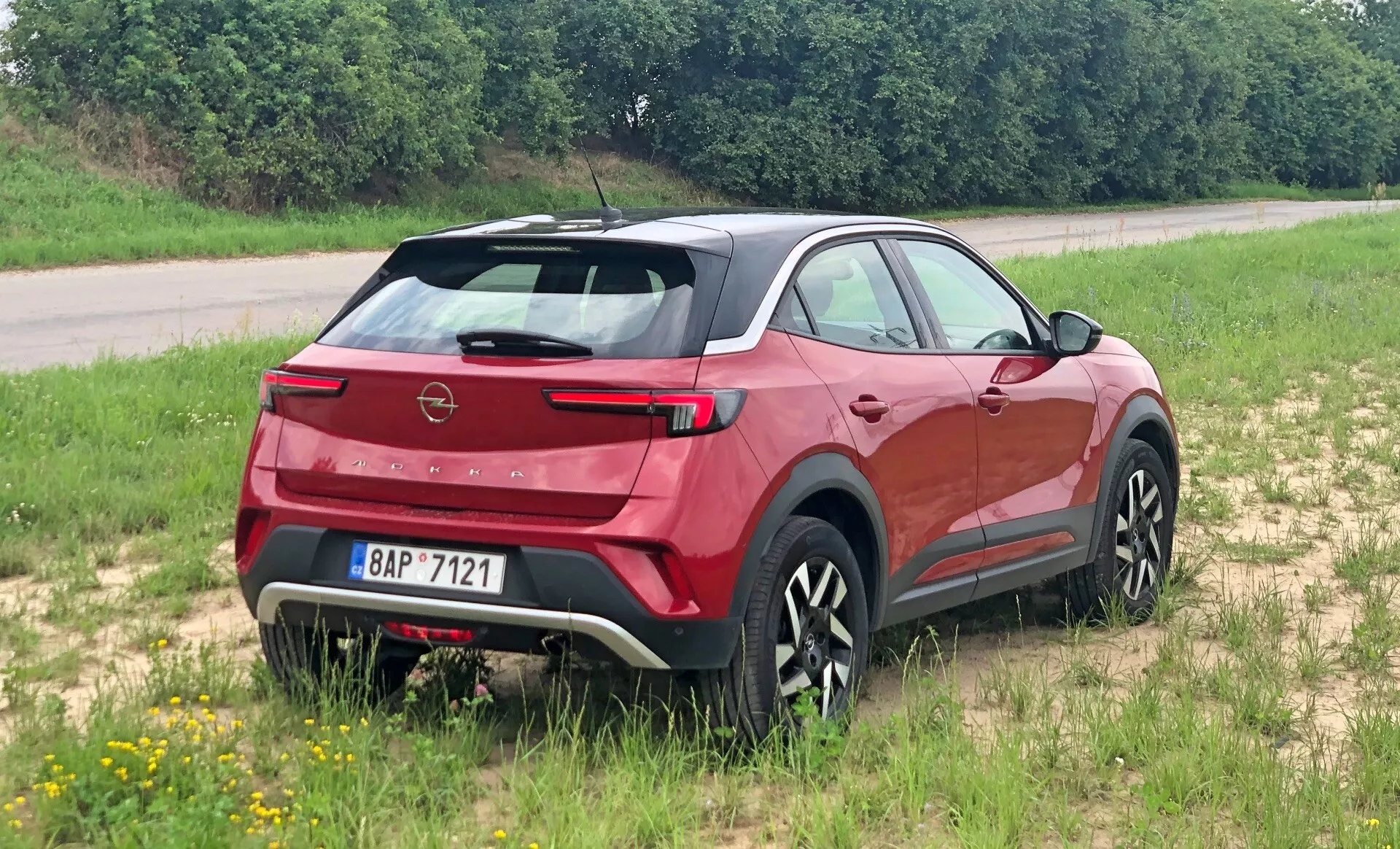 Moderno crossover SUV Opel Mokka 2021 visto da una prospettiva posteriore tre quarti, evidenziando lo spoiler posteriore, il parabrezza posteriore inclinato, il visibile logo del marchio e le eleganti luci posteriori. Posizionato su una superficie erbosa, mostrando il suo potenziale per il leggero fuoristrada. Il profilo laterale rivela linee scolpite della carrozzeria, rivestimenti neri e ruote dal design multirazze. Tergicristallo posteriore integrato, sensori di parcheggio e piastra skid di colore contrastante suggeriscono aerodinamica, praticità e robustezza.