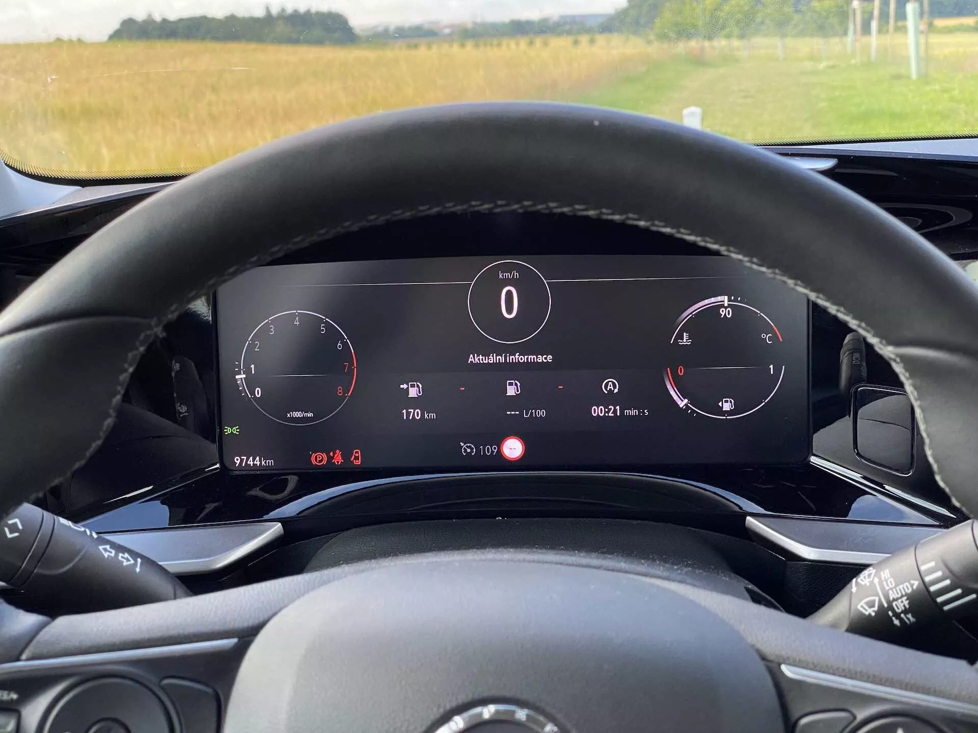 Vista interna della Opel Mokka 1.2 DI Turbo 2021 dalla prospettiva del conducente al volante. L'immagine mette in evidenza il cruscotto digitale dietro al volante che include tachimetro, contagiri e un pannello informativo che mostra la temperatura esterna (8°C), l'orologio, la posizione del cambio ('P' per parcheggio) e i dettagli del computer di bordo. Il volante presenta controlli multimediali visibili ed è parzialmente visibile. L'illuminazione ambientale suggerisce un contesto diurno con una vista rurale attraverso il parabrezza. Il veicolo è dotato di un cruscotto elettronico e caratteristiche tecnologiche avanzate.