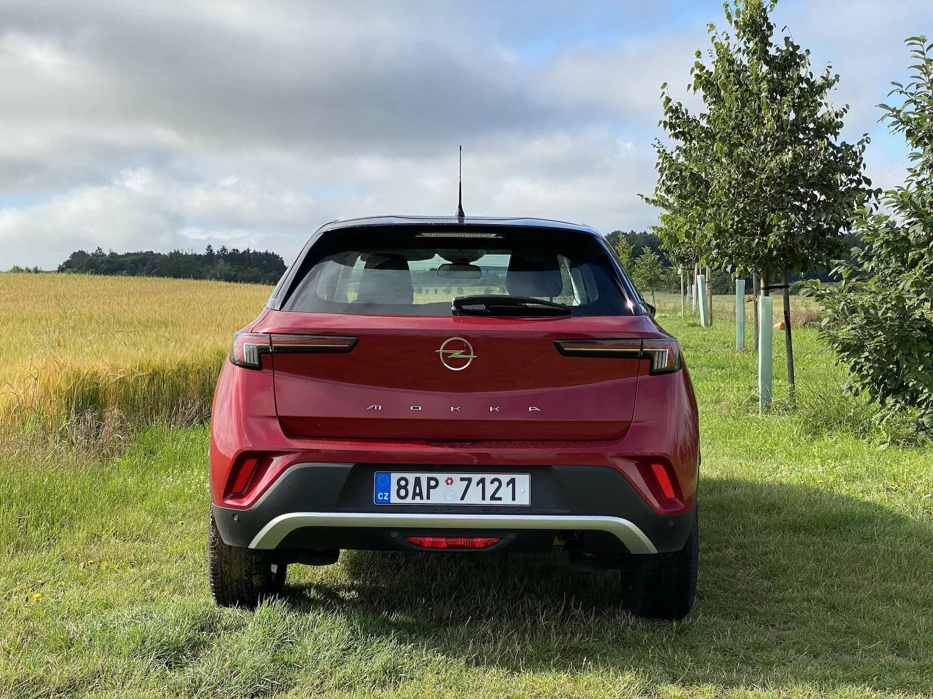 Vista posteriore di una Opel Mokka 1.2 DI Turbo 2021 rossa, che presenta un design moderno con linee affilate e il logo del produttore sul portellone, gruppi ottici posteriori a LED distintivi, paraurti posteriore con inserti a contrasto, sensori di parcheggio, riflettori integrati, tergilunotto, antenna a pinna di squalo, spoiler posteriore e targa in un ambiente rurale con erba, campo coltivato e alberi.