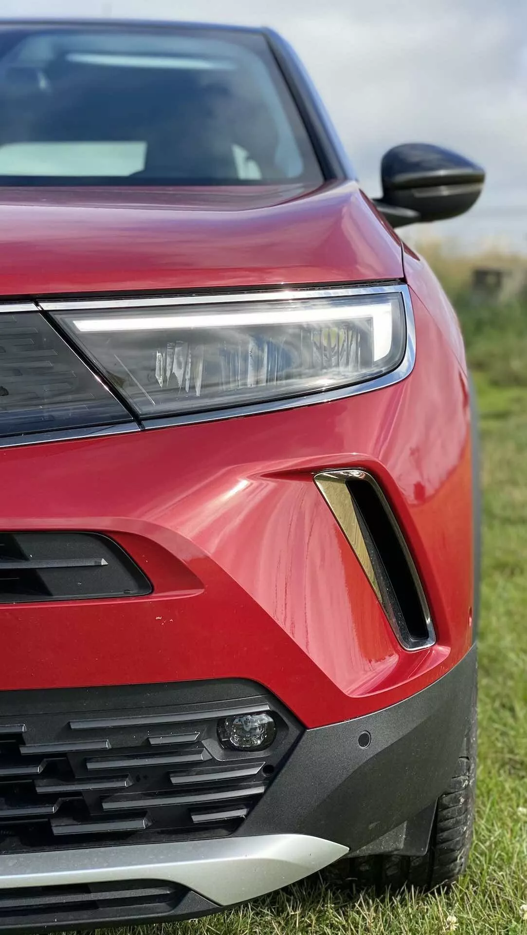 Vista ravvicinata e angolata del lato anteriore sinistro di una Opel Mokka 2021 rossa. La foto cattura il faro con illuminazione LED o HID, parte della griglia con lamelle orizzontali, l'alloggiamento del fendinebbia con contorno nero e argento, vernice carrozzeria rosso brillante con contorni angolari audaci, specchietto laterale aerodinamico e cofano e parafango parzialmente visibili. L'auto è parcheggiata sull'erba con uno sfondo sfocato.