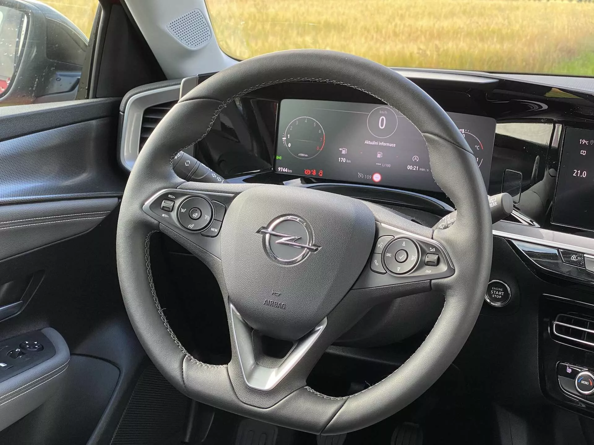 Vista interna della Opel Mokka 2021 dalla prospettiva del sedile passeggero anteriore, che mostra il volante con pulsanti di controllo audio e cruise control, quadro strumenti digitale che visualizza tachimetro e altre metriche, e console centrale con sistema di infotainment touchscreen.