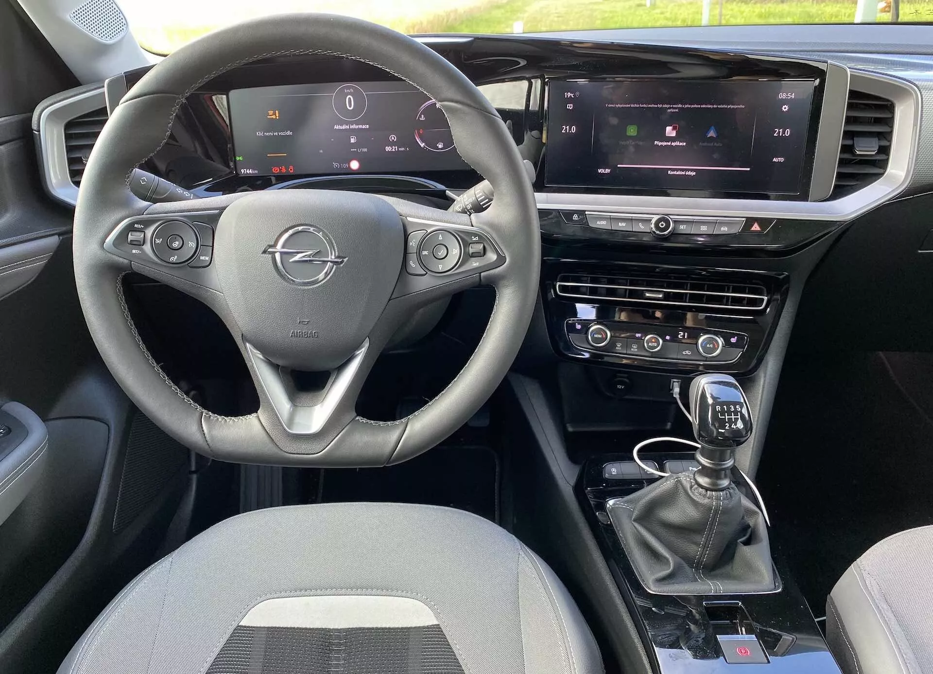 Interni della Opel Mokka 2021 dalla prospettiva del passeggero anteriore, con volante a tre razze con comandi integrati, cruscotto completamente digitale, touch screen centrale di infotainment con controllo del clima e navigazione e leva del cambio manuale. La disposizione include sistemi di controllo doppi e ulteriori pulsanti per modalità di guida, con tappezzeria in tessuto.