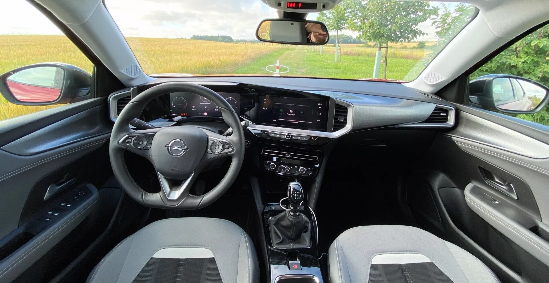 Vista interna dal sedile posteriore di un Opel Mokka 2021 1.2 DI Turbo. Dotato di volante multifunzione a tre razze, display digitale di tachimetro e contagiri dietro al volante, schermo touchscreen infotaiment in verticale, interruttori per il controllo del clima, cambio manuale con portabicchieri, sedili anteriori sportivi bicolore, pannelli porte abbinati con bracciolo, specchietto retrovisore visibile, finestrini parzialmente abbassati e comandi aggiuntivi per tergicristalli e indicatori di direzione.