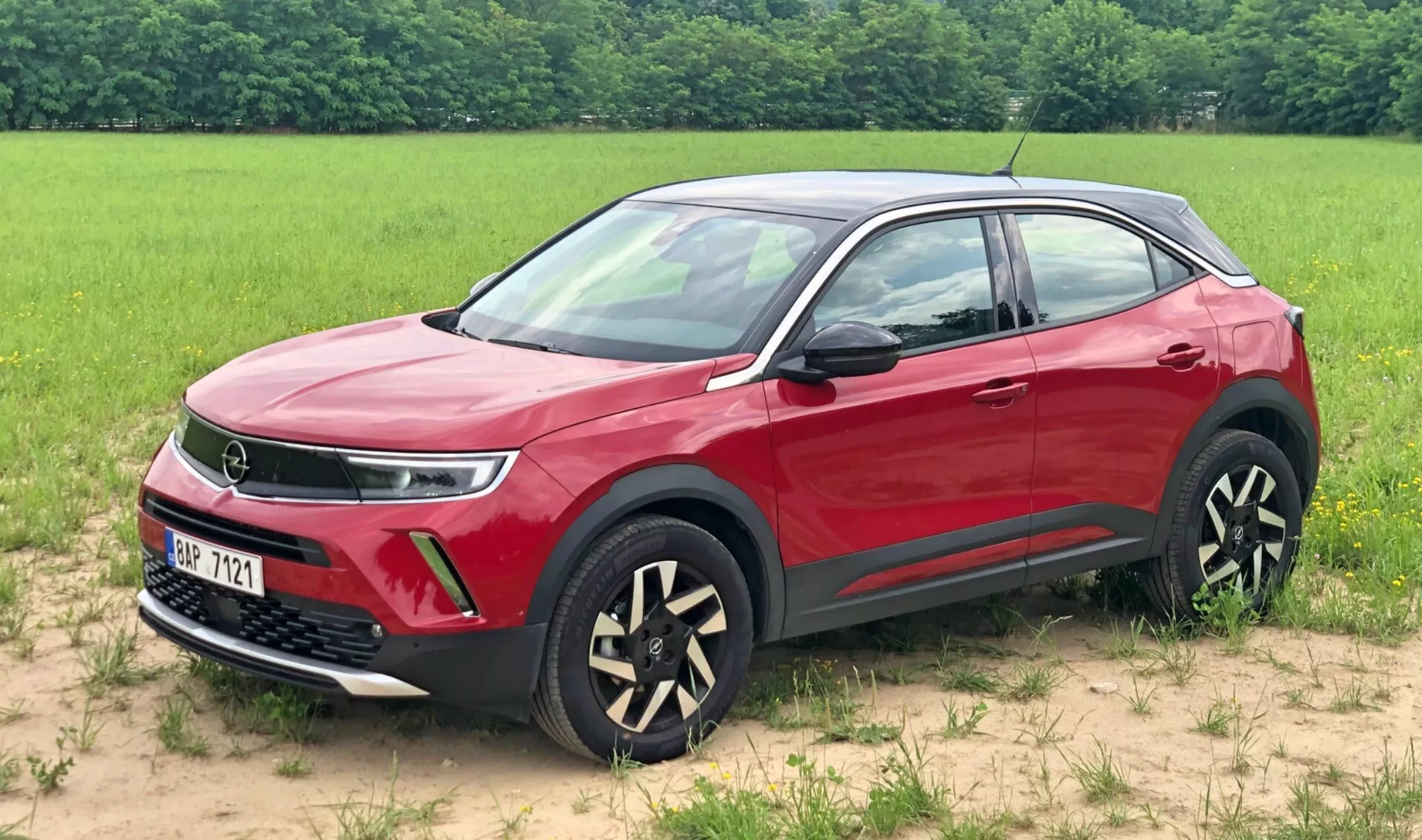 Immagine di un SUV Opel Mokka 2021 rosso parcheggiato su una superficie erbosa con un campo sullo sfondo. Questa vista anteriore a tre quarti mette in evidenza la griglia, indicando potenzialmente una tecnologia avanzata del gruppo propulsore, eleganti luci diurne a LED, i fendinebbia e cerchi in lega bicolori unici. Anche gli specchietti e le maniglie delle porte in tinta con la carrozzeria, la linea delle spalle dinamica, il rivestimento inferiore nero protettivo e le cornici cromate dei finestrini sono visibili, mostrando le caratteristiche di design moderni e lussuosi del veicolo.
