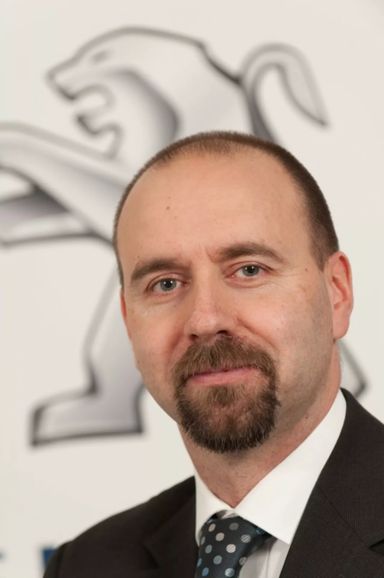 Ritratto professionale di un uomo in abito e cravatta con il logo del leone Peugeot sullo sfondo, nessuna auto visibile