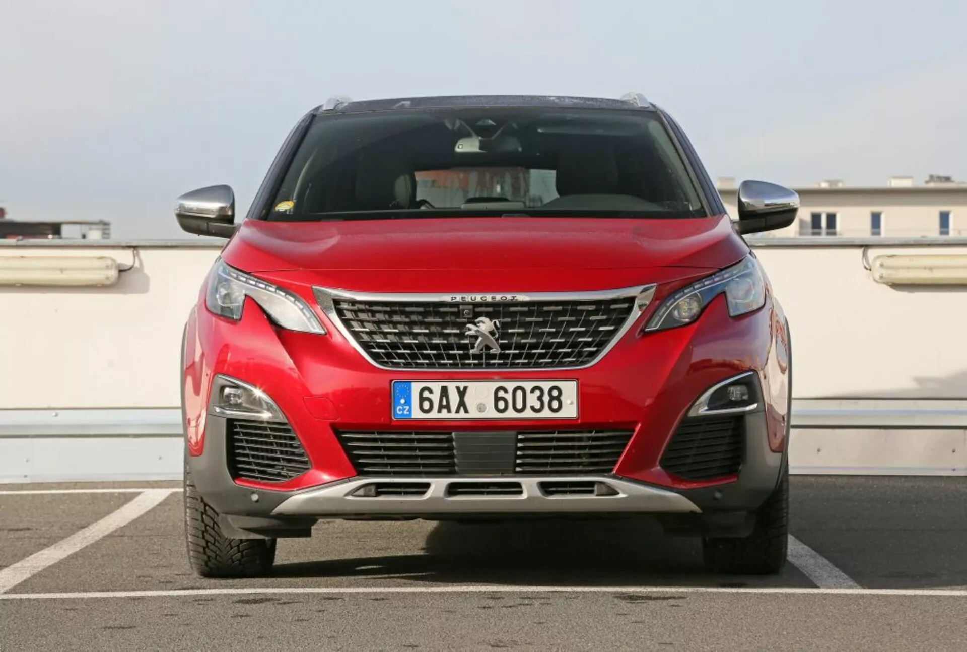 Vista frontale di una Peugeot 3008 rossa del 2016 che mostra l'emblema sulla griglia, fari angolari, prese d'aria inferiori con luci antinebbia, accenno dei contorni del cofano su uno sfondo urbano con cielo limpido