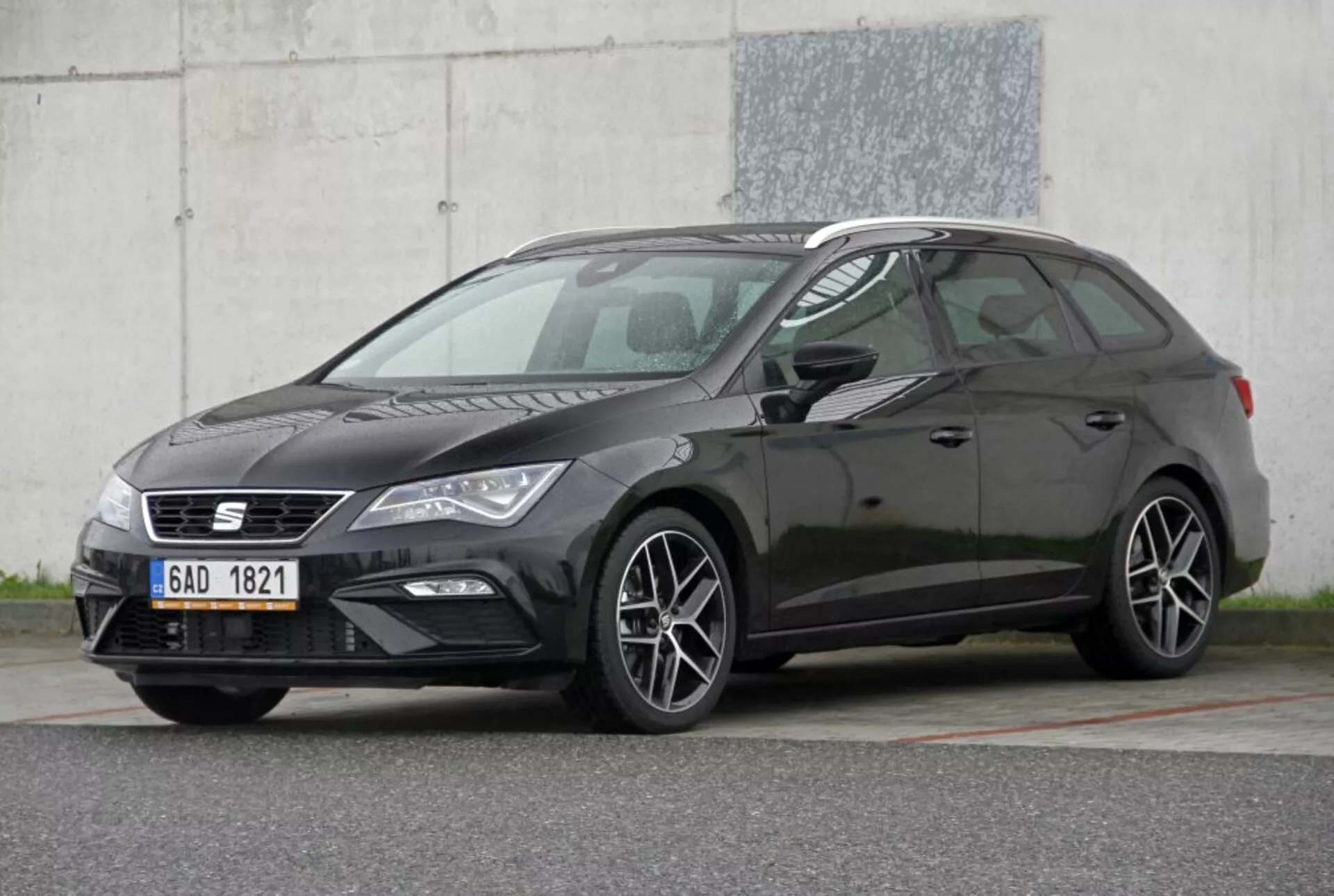 Veicolo hatchback Peugeot 308 SW di colore scuro del 2013 che mostra le sue caratteristiche: griglia stretta con emblema, unità fari eleganti con luci diurne, ruote in lega a multi-razi che suggeriscono un modello o livello di allestimento sportivo, profilo laterale distintivo da hatchback con la linea del tetto discendente, in un contrastante colore scuro della carrozzeria, ripreso da un angolo frontale di tre quarti contro uno sfondo industriale