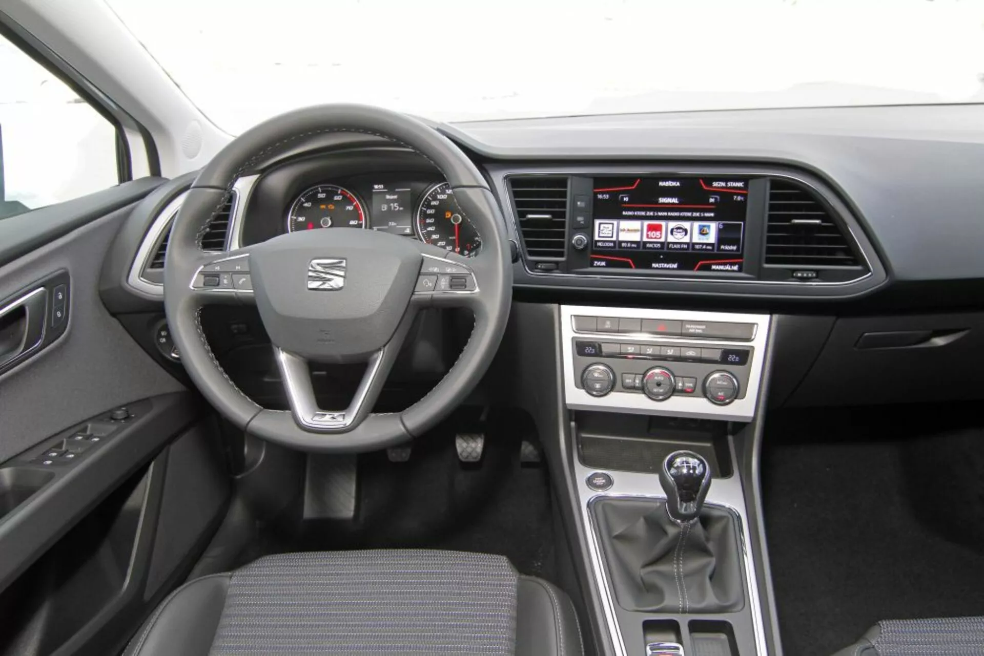 Vista interna di una Peugeot 308 SW del 2013 che mostra il volante con logo e comandi multifunzione, tachimetro e contagiri analogici con display digitale, sistema di infotainment touch sul cruscotto, pulsanti di controllo manuale del clima sulla console centrale, leva del cambio manuale e tappezzeria pratica in tessuto scuro.