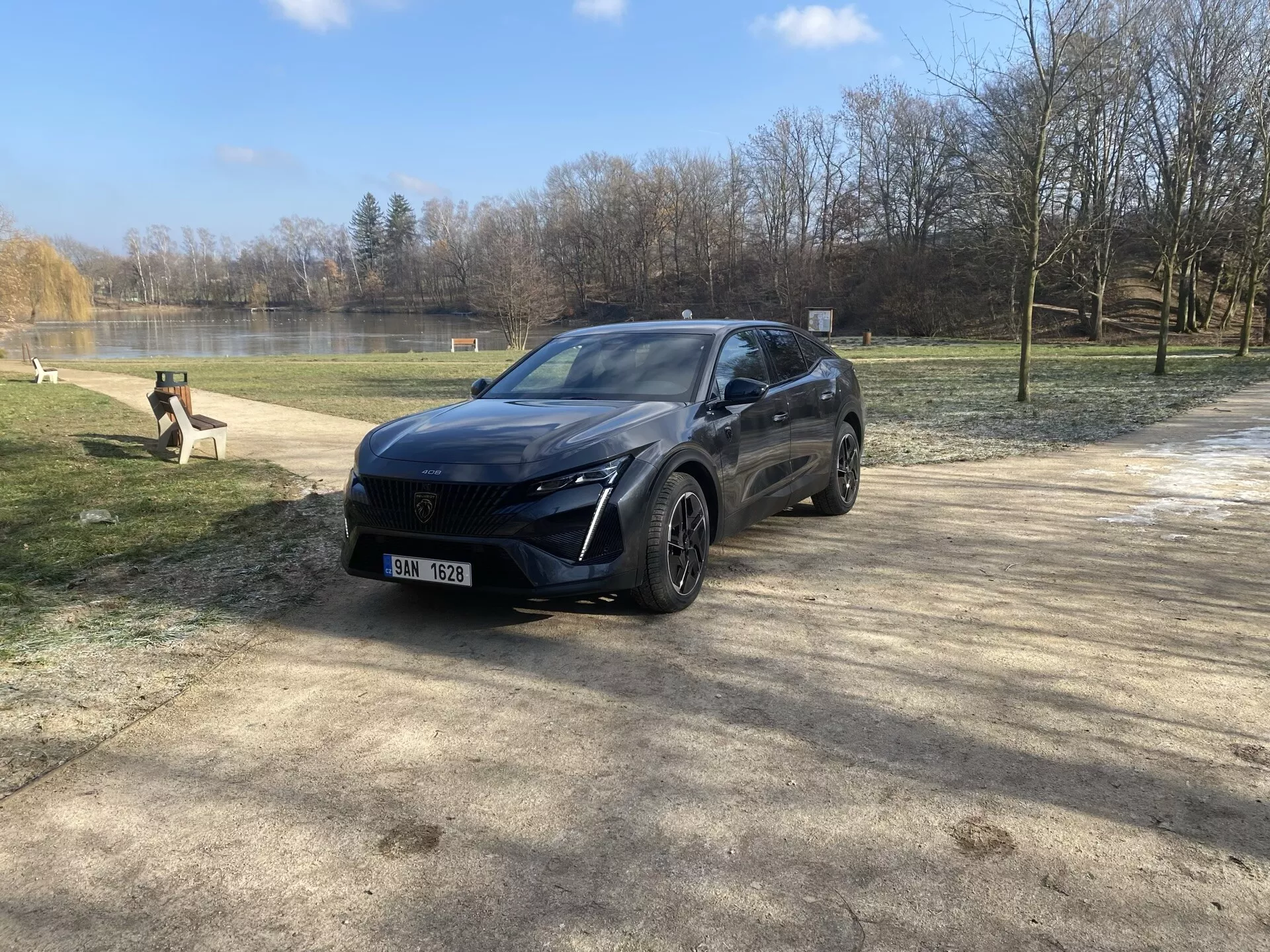 Peugeot 408 del 2023 parcheggiata in un ambiente naturale durante l'inverno o l'inizio della primavera, che mette in mostra il suo design moderno da crossover, fari LED, sistema di sensori frontali, cerchi in lega e finitura premium da una vista angolare frontale di tre quarti sotto la luce del sole con flare dell'obiettivo.