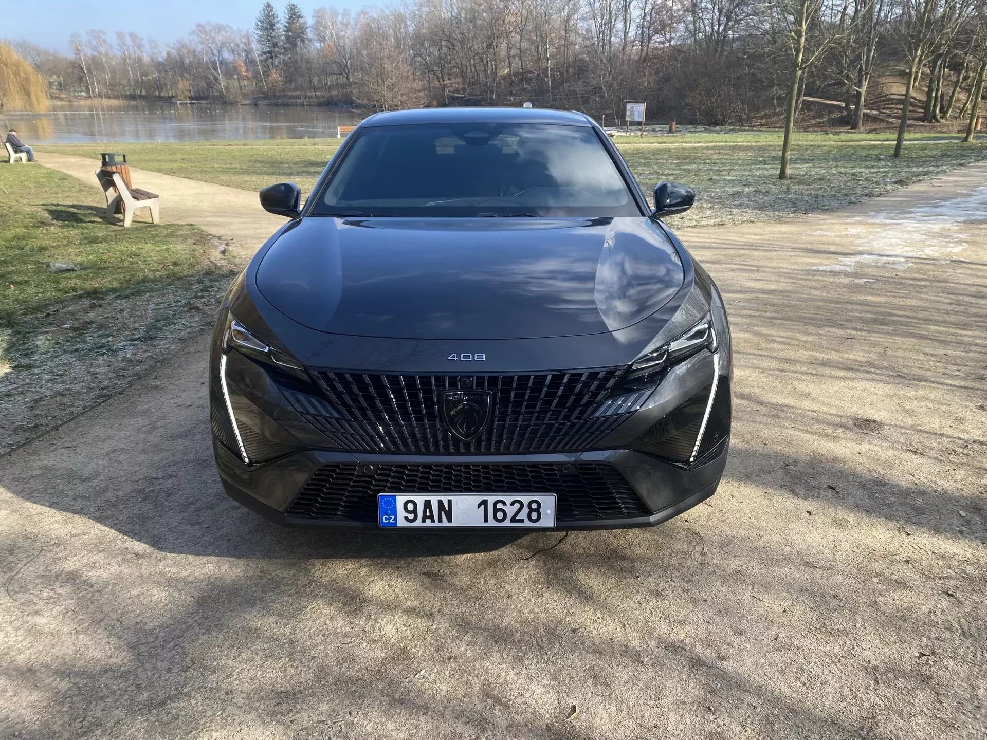 Vista frontale della Peugeot 408 2023 parcheggiata su una superficie ghiaiosa con alberi e uno specchio d'acqua sullo sfondo, evidenziando la sua grande griglia con motivi a forma di diamante, fari LED eleganti, linee scolpite sul cofano e funzionalità avanzate di assistenza al guidatore