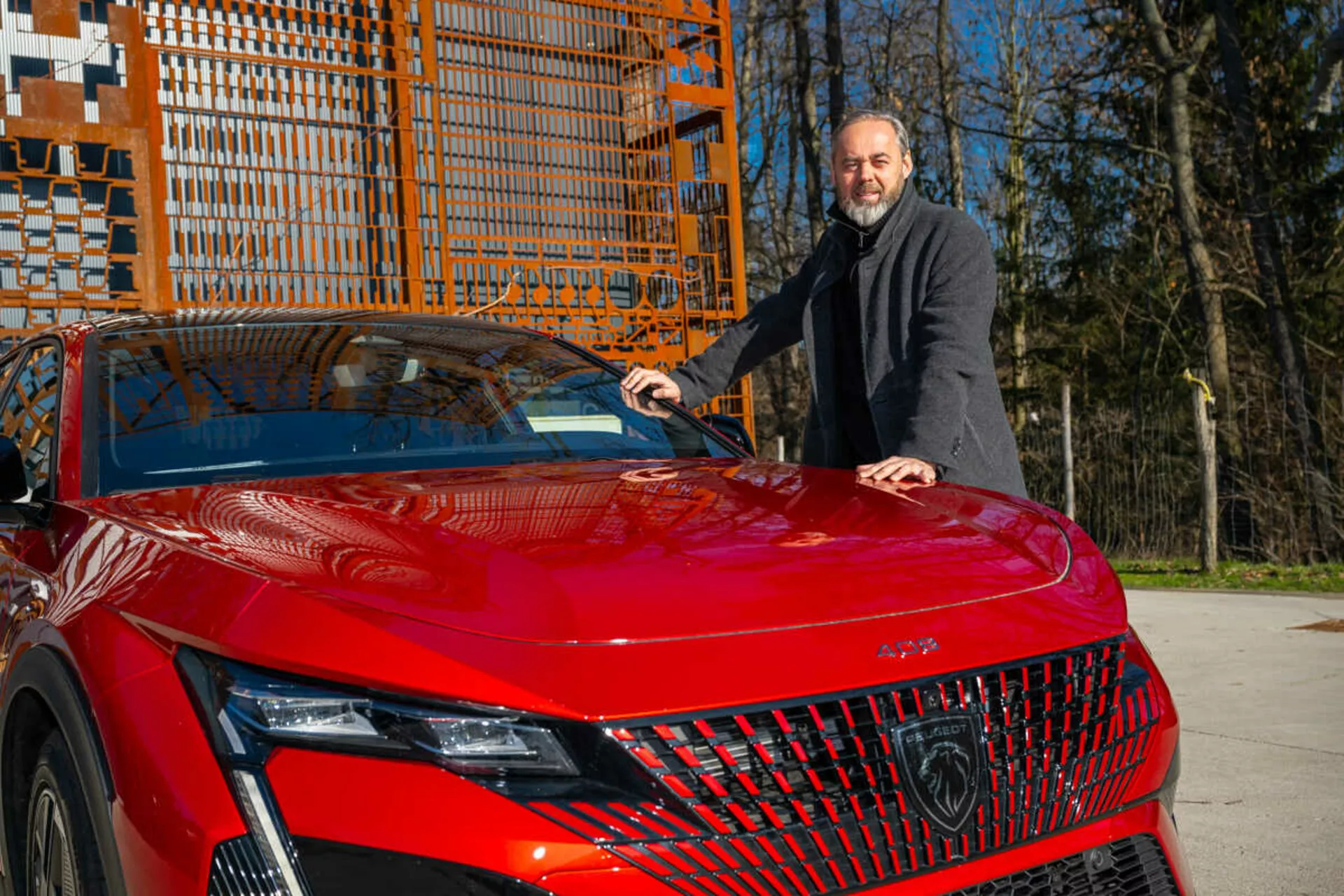 Modello rosso della Peugeot 408 del 2023 che mostra il suo quarto anteriore sinistro alla luce del giorno, evidenziando i fari LED dettagliati, la griglia geometrica e il cofano sportivo e aerodinamico con un uomo in un cappotto scuro accanto ad esso che pone la mano sul cofano
