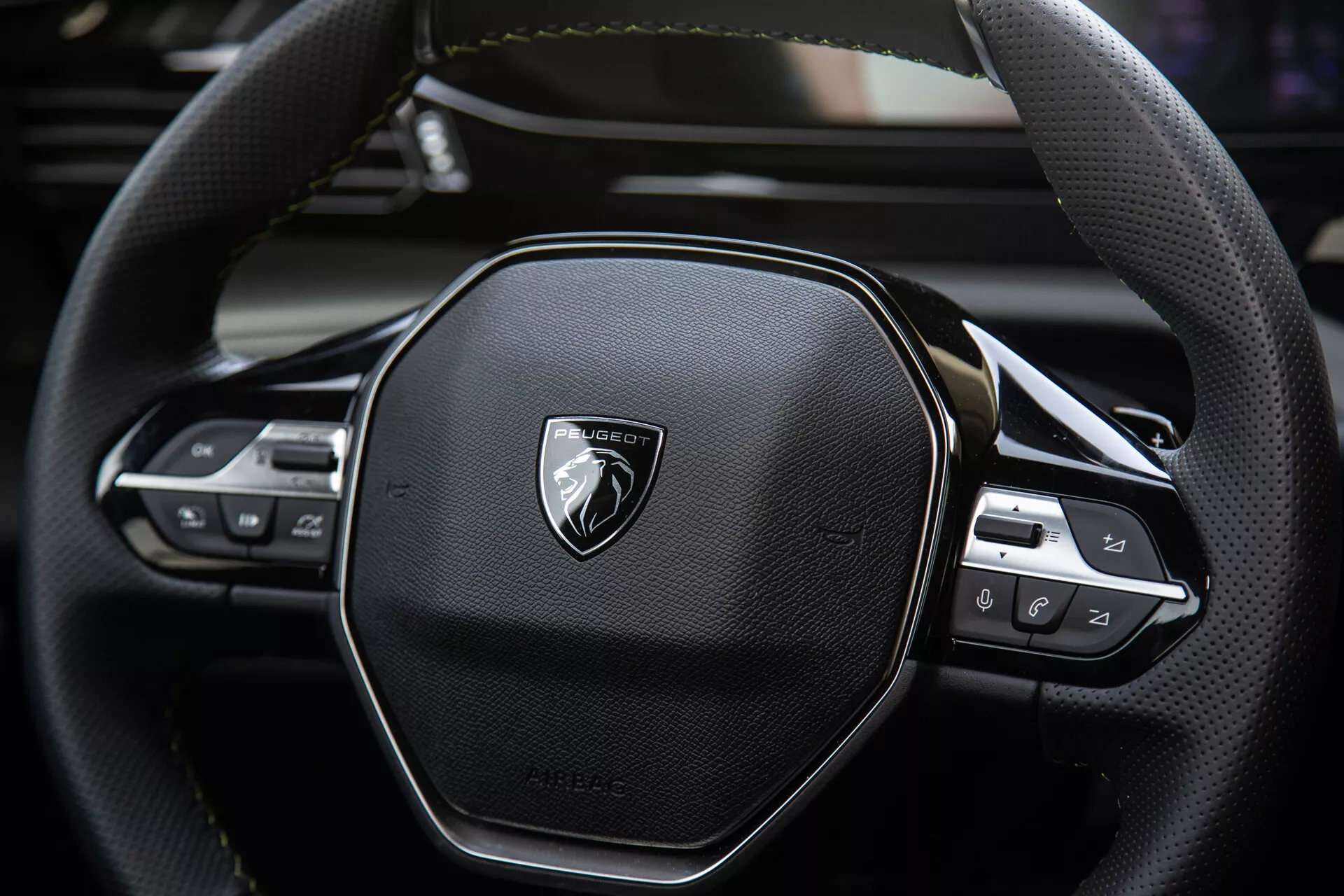 Vista ravvicinata del volante del Peugeot 408 2023 che mostra design ergonomico esagonale, cuciture in pelle, controlli tecnologici integrati per audio e impostazioni di crociera, con emblema su metallo spazzolato.