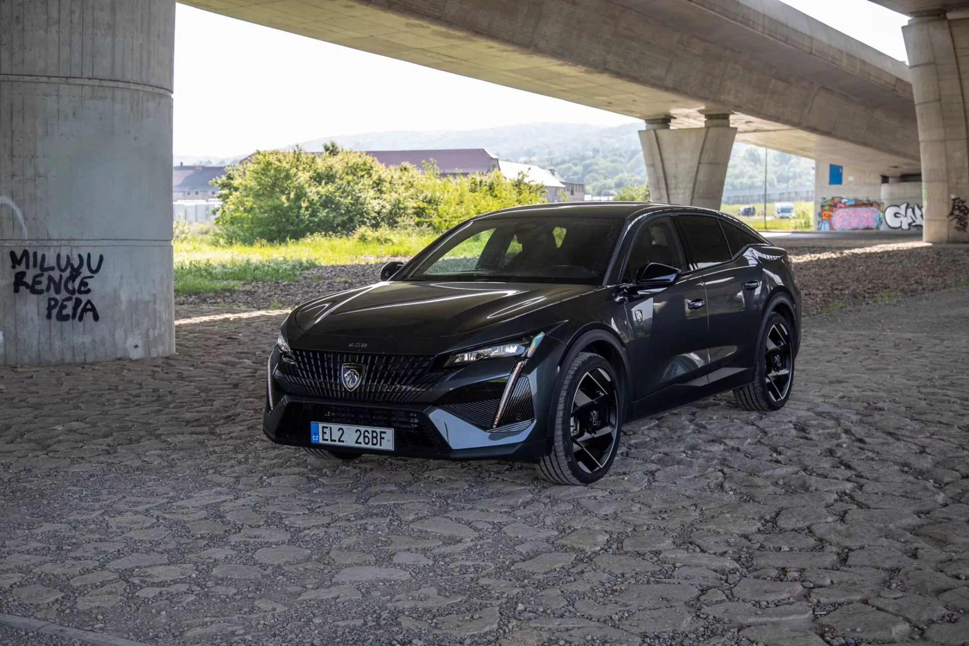 Peugeot 408 2023 fotografato da un angolo di tre quarti che evidenzia i suoi fari LED eleganti, griglia sagomata e paraurti anteriore sportivo aerodinamico sotto luce naturale diurna in un'area pavimentata sotto un cavalcavia
