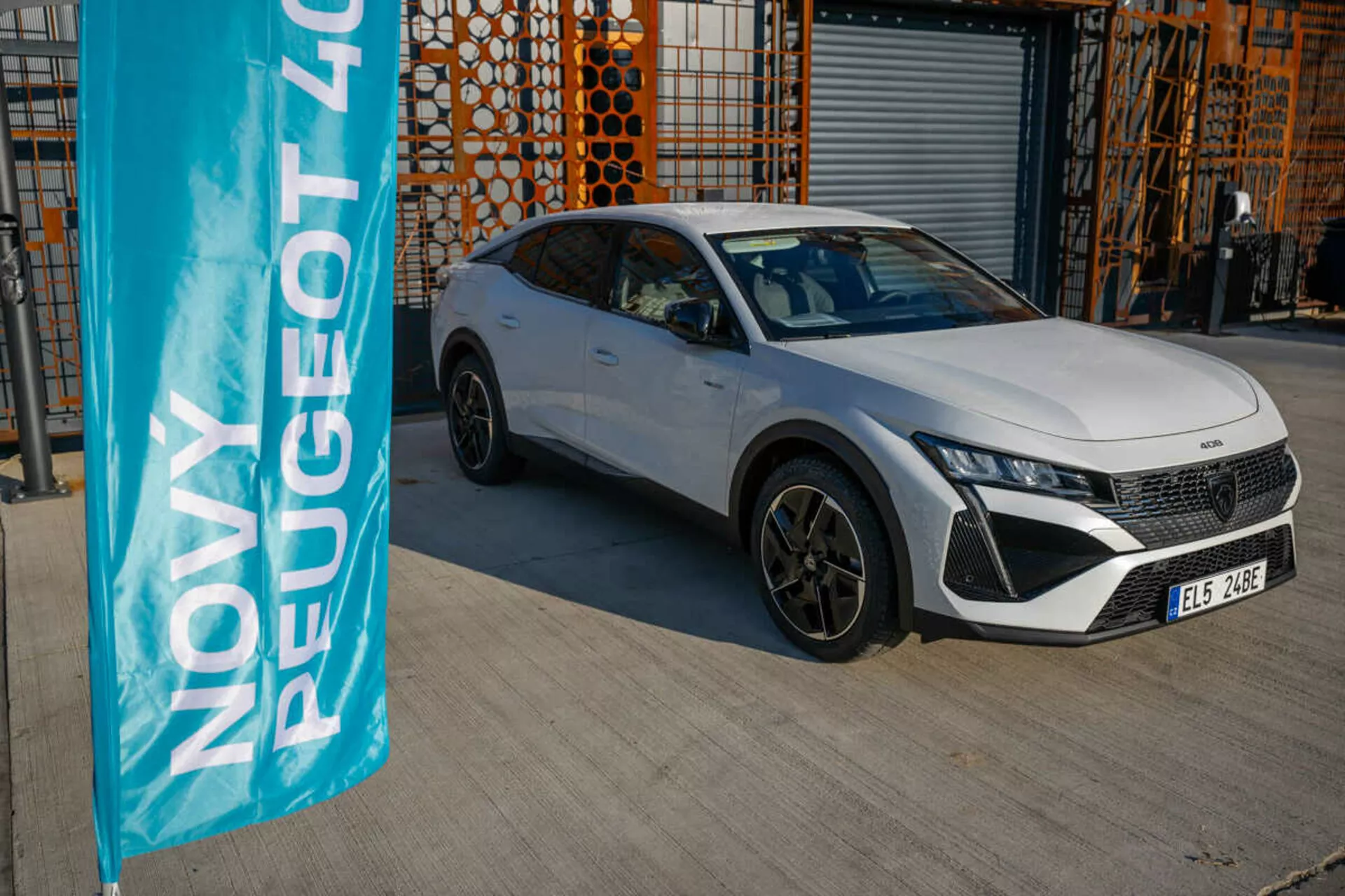 Peugeot 408 bianca del 2023 parcheggiata all'aperto con architettura industriale sullo sfondo, che mostra le caratteristiche di design e tecnologia del veicolo alla luce naturale del giorno