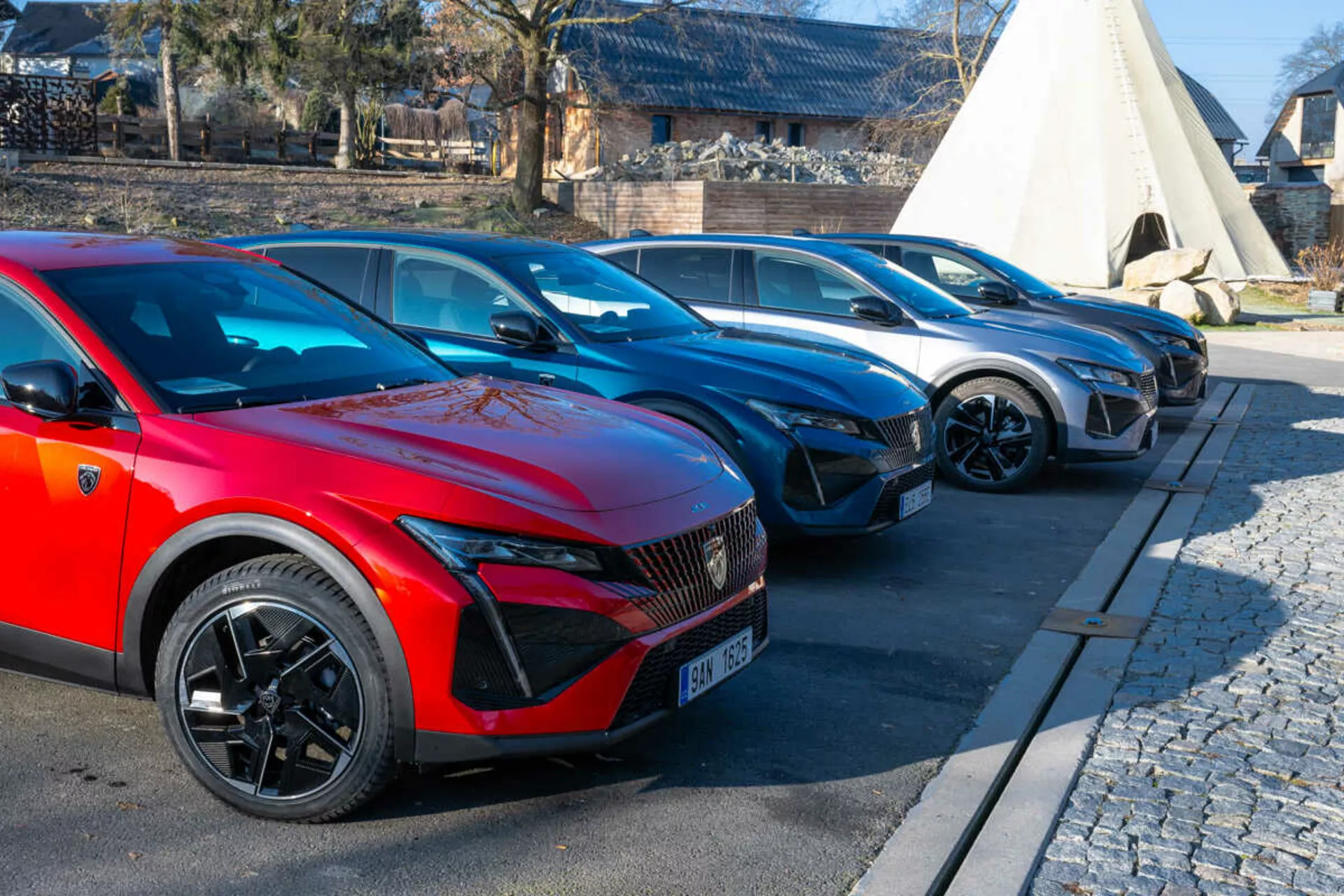 Lineup Peugeot 408 del 2023 con auto rossa in primo piano con ruote sportive a più razze e fari LED indicativi di modelli elettrici o ibridi, parcheggiata su area pavimentata accanto a selciato