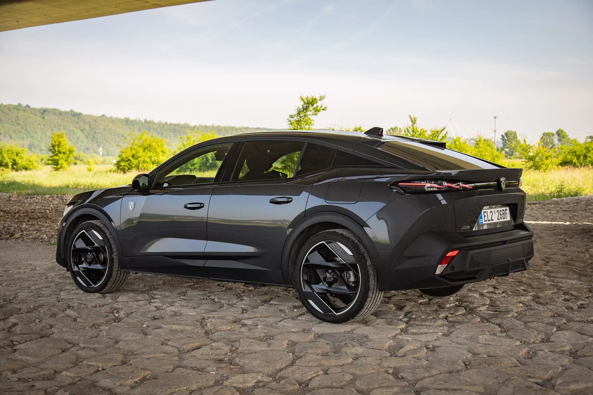 SUV crossover Peugeot 408 del 2023 in finitura scura, catturando una visuale posteriore di tre quarti che evidenzia un design aerodinamico con angoli marcati, tetto sportivo inclinato, grandi ruote in lega nere, fanali posteriori LED distintivi e uno spoiler posteriore sottile su una superficie in ciottoli
