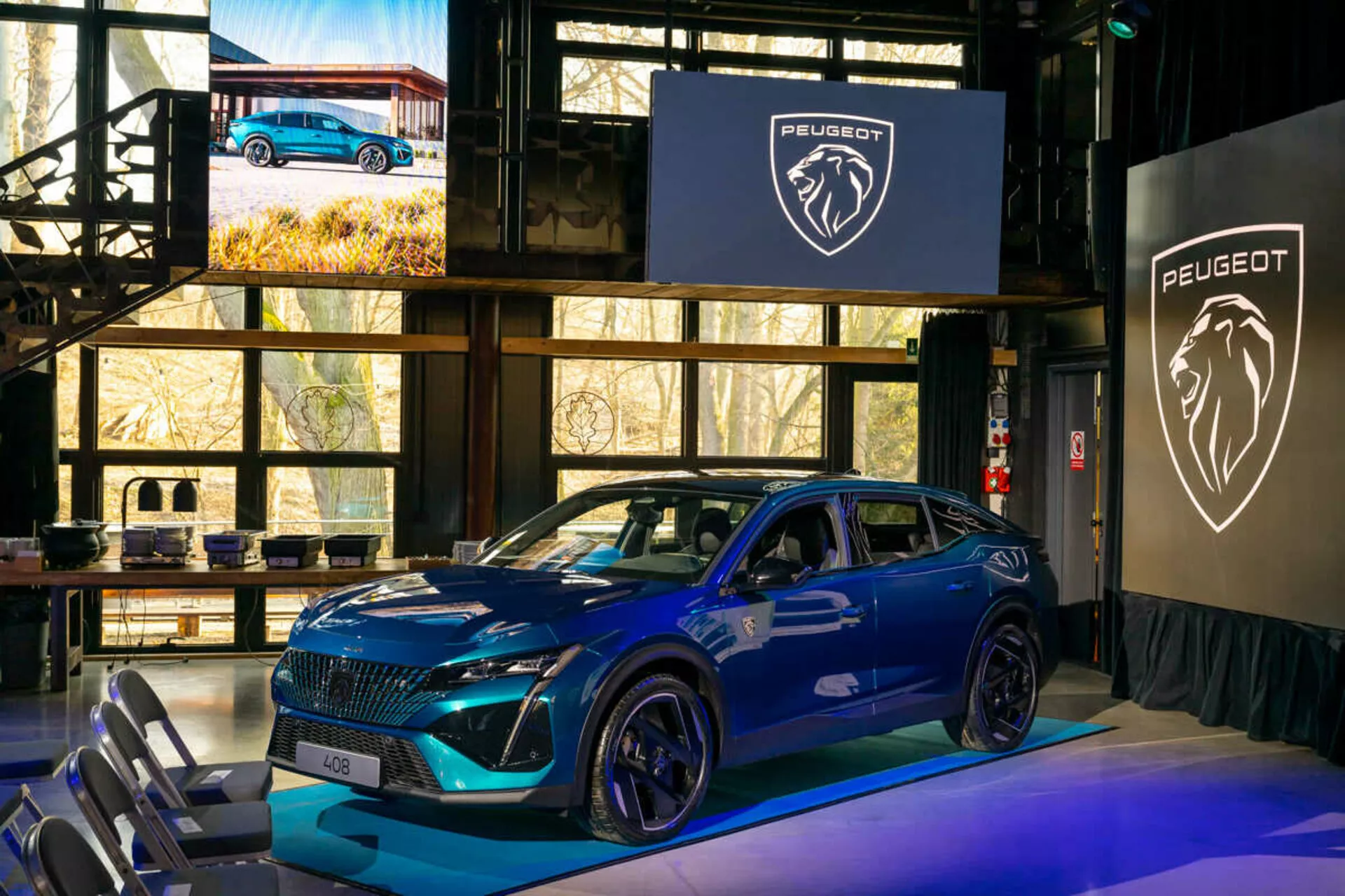 Peugeot 408 blu del 2023 esposta in un evento indoor, caratterizzata da una griglia distintiva, fari LED affilati, cofano sportivo sagomato con prese d'aria, grandi ruote in lega e un profilo laterale dinamico con indicatori di direzione integrati sugli specchietti laterali montati sulla porta