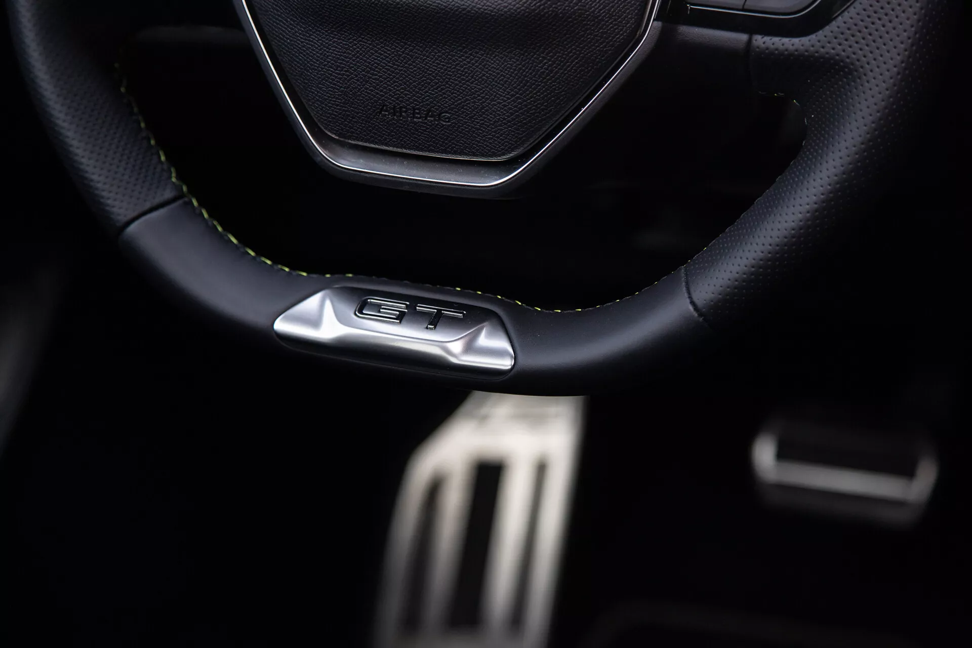 Interno della Peugeot 408 2023 con focus sul volante sportivo in pelle con cuciture e emblema GT, pedali metallici ad alte prestazioni e dettagli premium del cockpit scuro