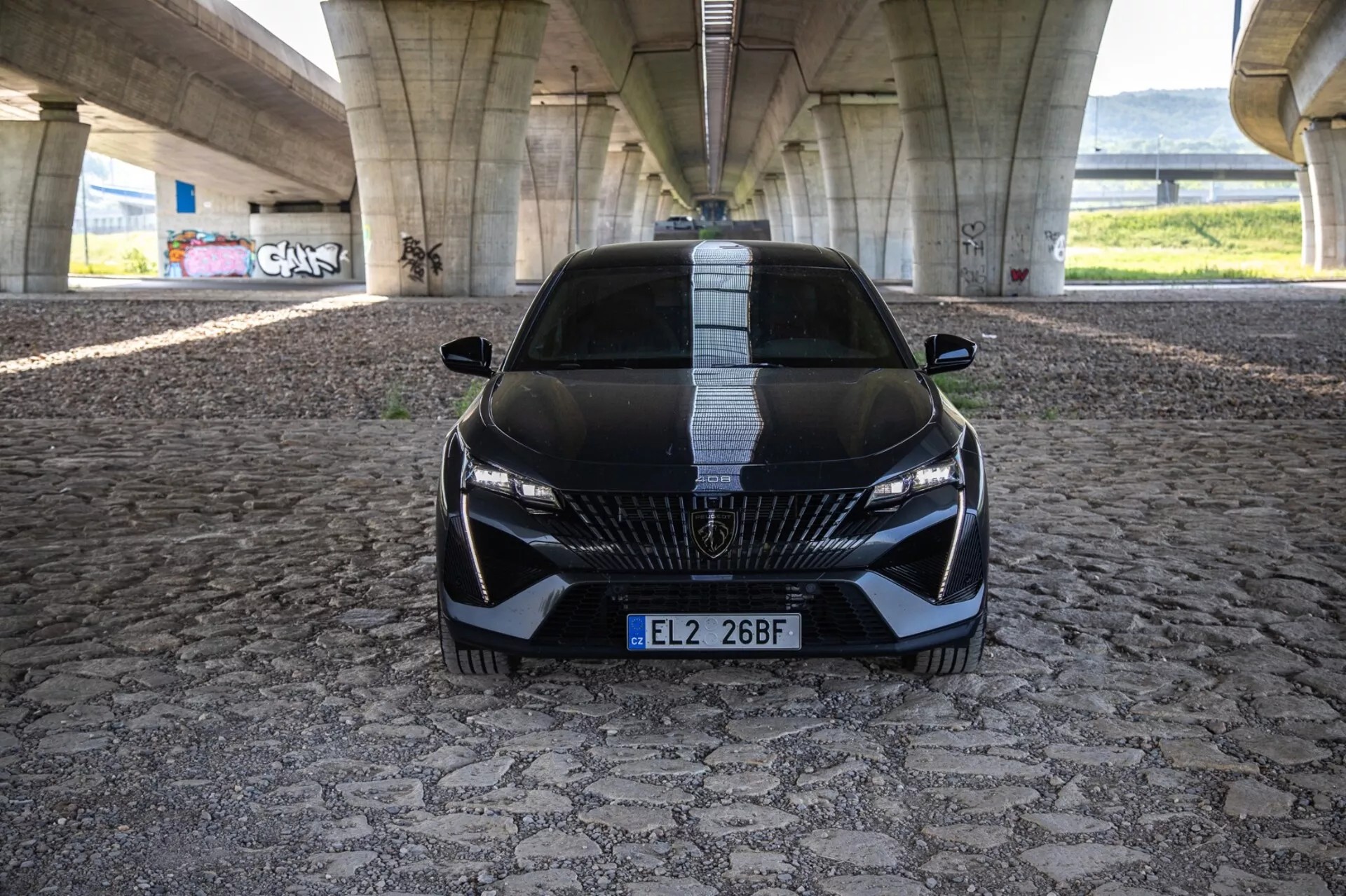 Peugeot 408 del 2023 in una vista frontale dinamica che mette in evidenza la sua postura aggressiva e il design sportivo con griglia dettagliata, fari LED eleganti e caratteristiche aerodinamiche su un pavimento decorato sotto un ponte urbano