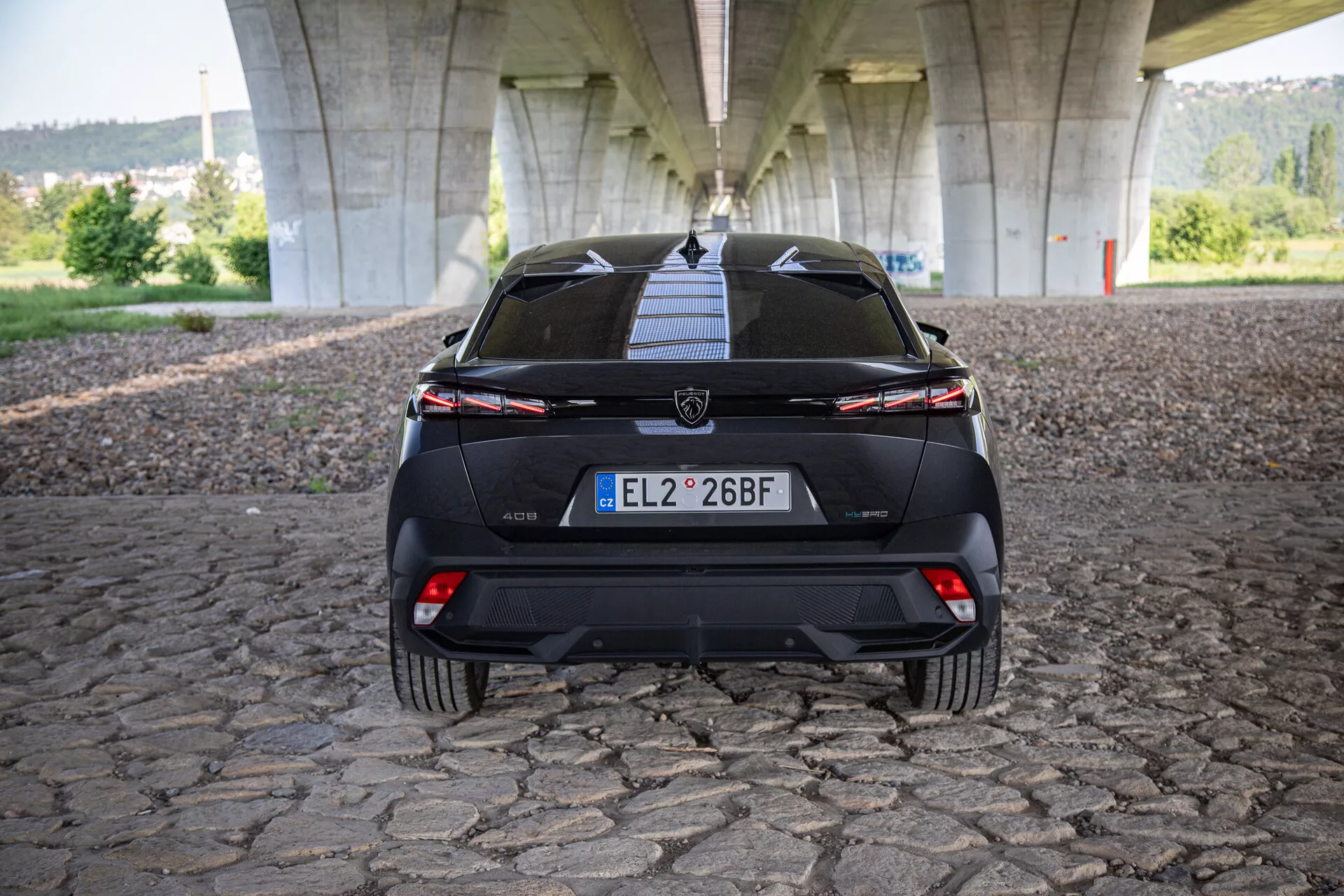 Vista posteriore della Peugeot 408 2023 che mette in mostra portellone, paraurti, diffusore inferiore, luci posteriori con una barra luminosa connessa e sensori di assistenza al parcheggio, fotografata sotto un ponte su superficie in ciottoli