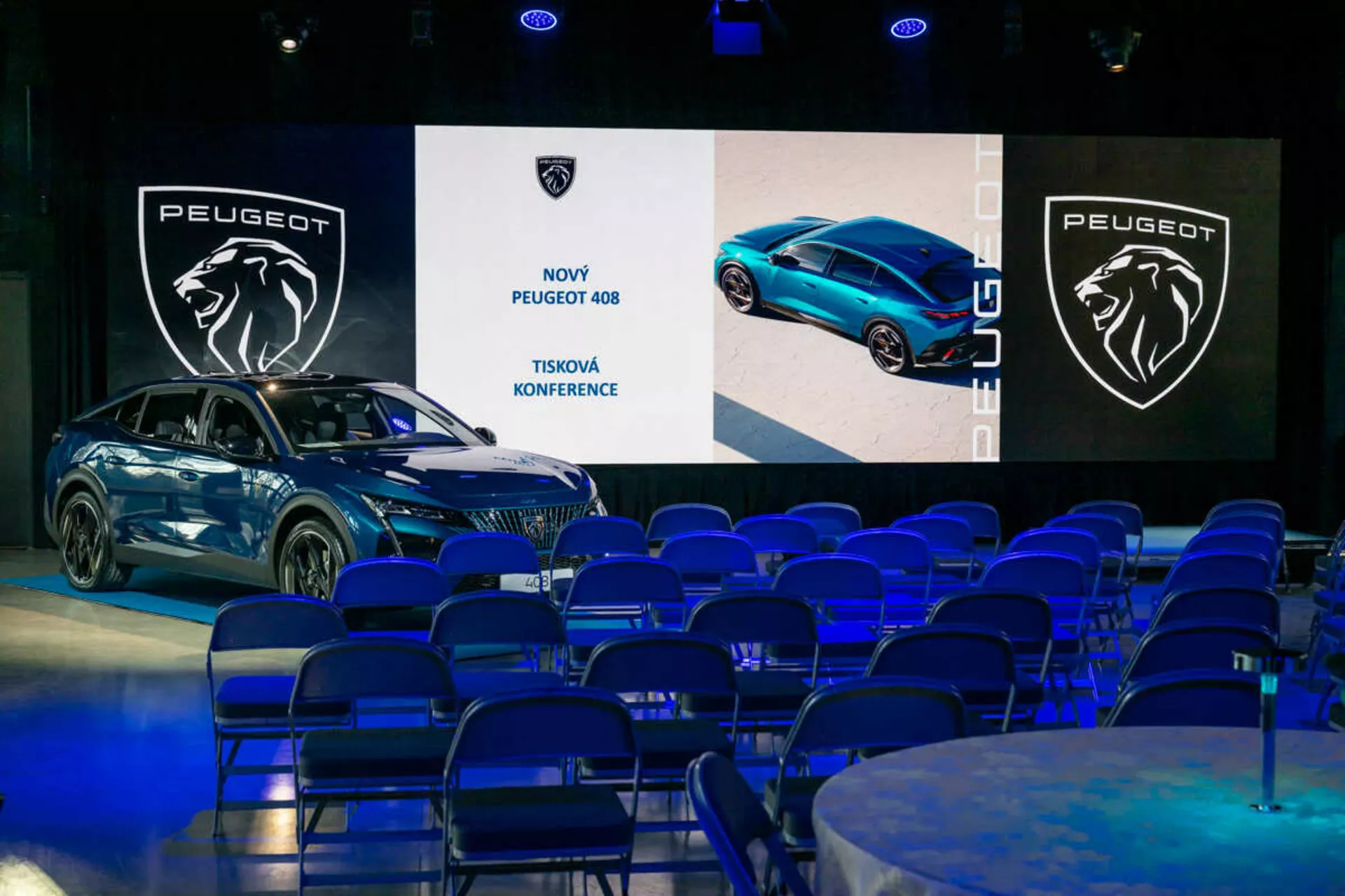 Peugeot 408 esposta a una conferenza stampa, vista in un angolo di 45 gradi che evidenzia i suoi profili frontale e laterale, il design elegante e i fari LED, con schermi digitali e branding sullo sfondo