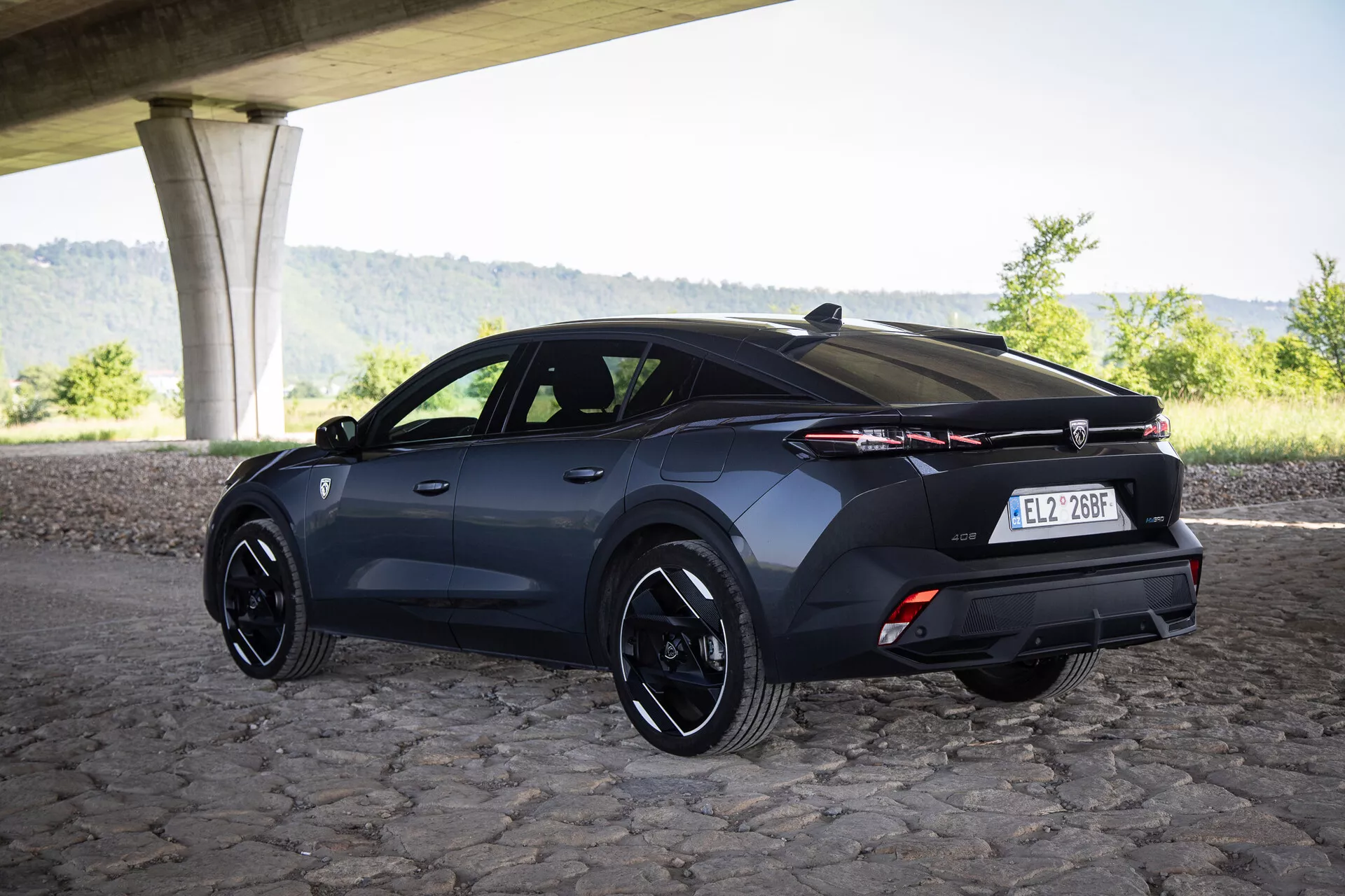 Vista posteriore tre quarti di una Peugeot 408 hatchback 2023 sotto un ponte su pavimentazione in sampietrini con cerchi sportivi e tecnologia di illuminazione avanzata