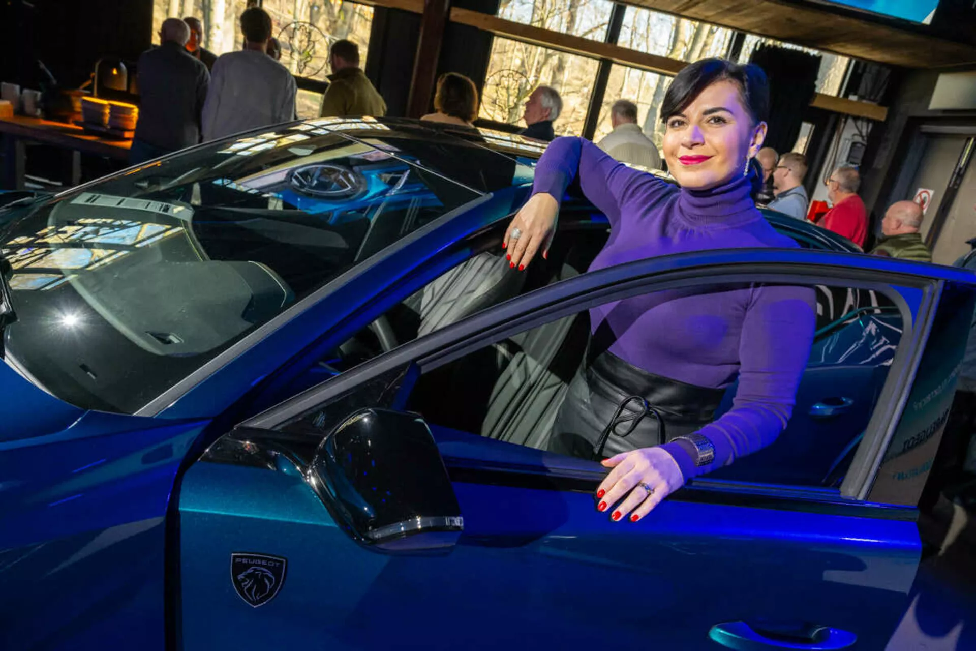 Auto sportiva blu vibrante, possibilmente una Lamborghini, catturata da un angolo di tre quarti che enfatizza le sue linee eleganti, fari LED affilati e iconico logo a forma di scudo, con una persona che interagisce casualmente con il veicolo suggerendo lusso e stile.