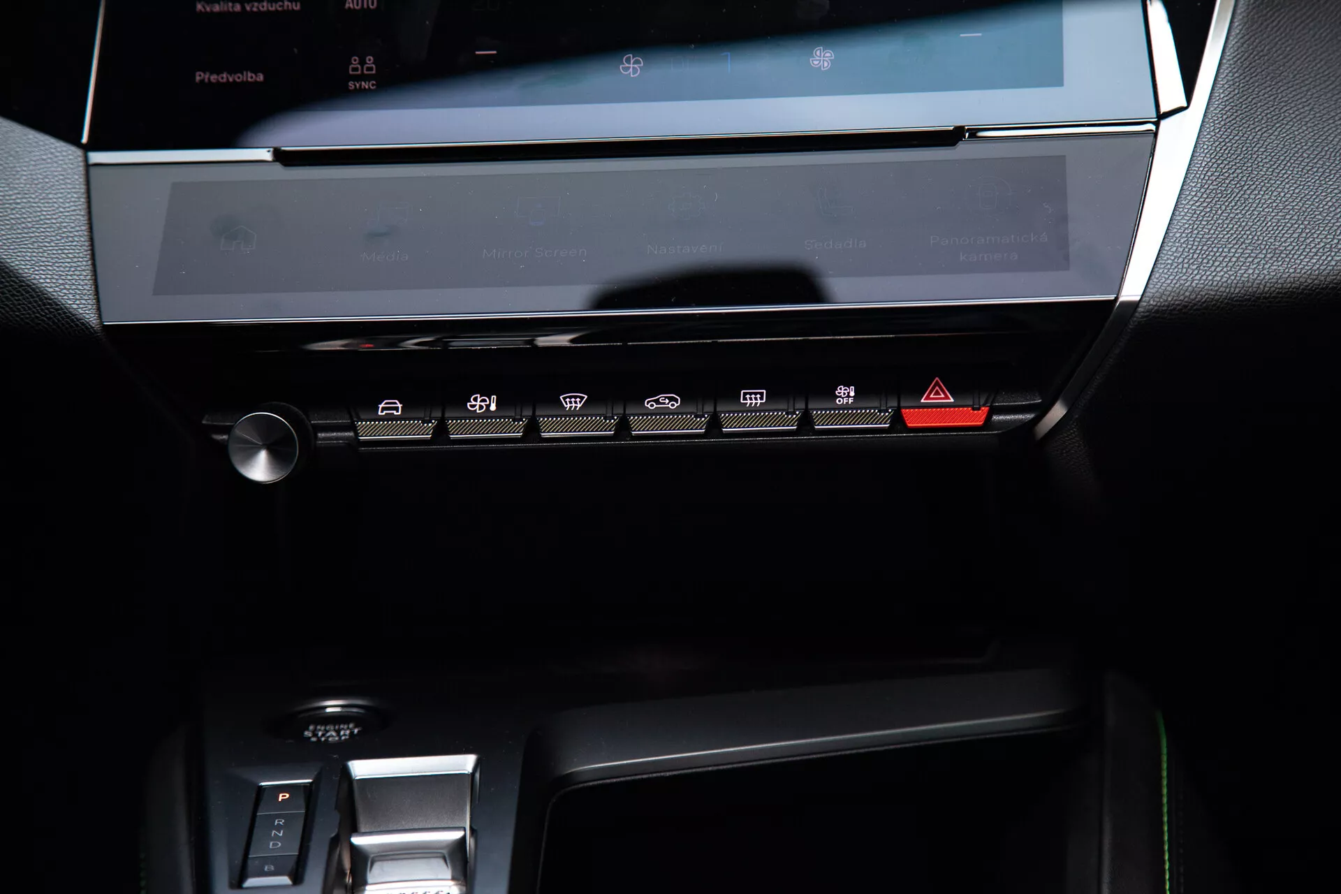 Cruscotto centrale del Peugeot 408 del 2023 con sistema di infotainment touchscreen spento, pannello di controllo del clima con pulsanti touch-sensitive e parte dell'area del cambio elettronico, illuminato da una luce ambiente soffusa che ne evidenzia il design moderno ed elegante con tonalità scure ed accenti metallici.