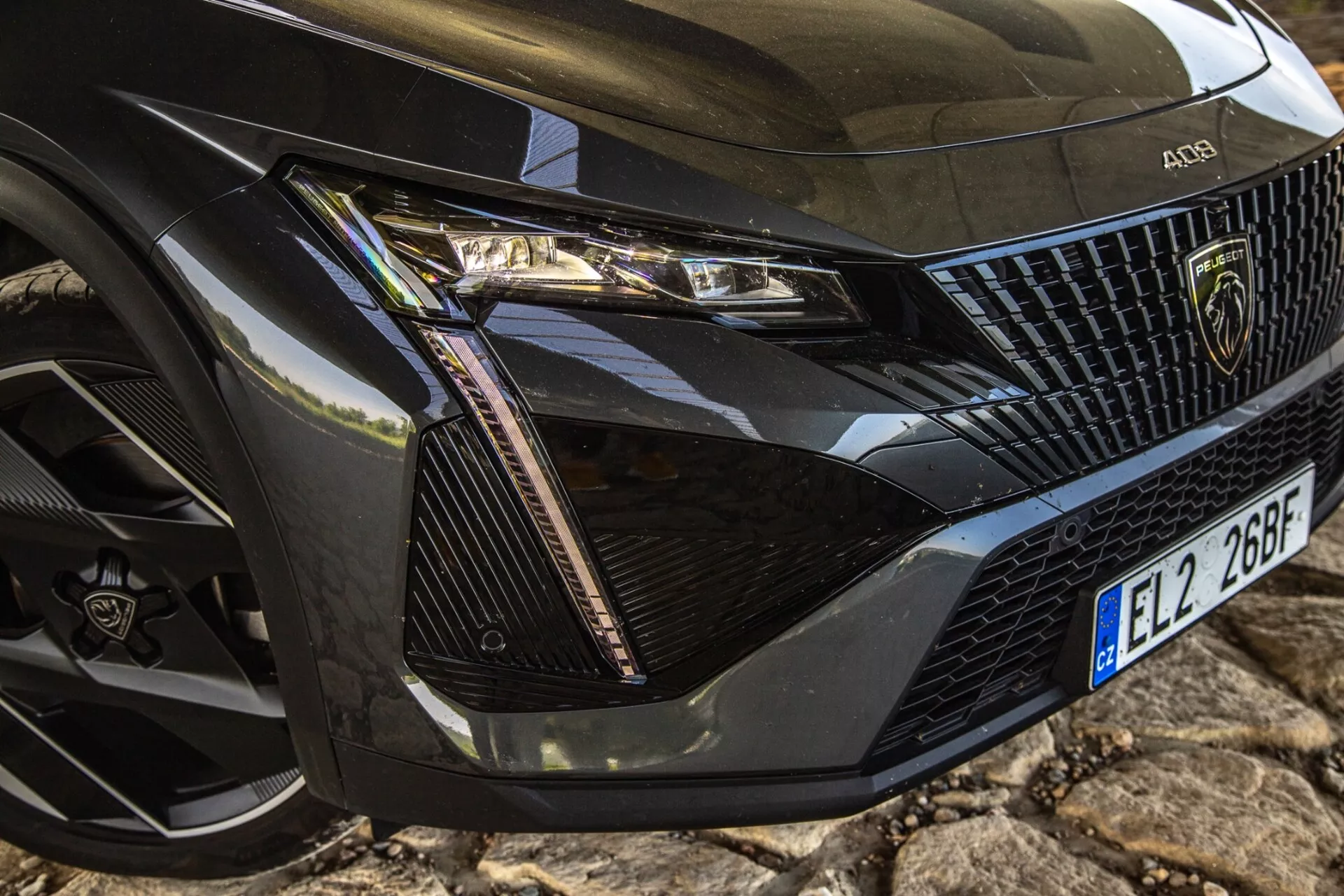Primo piano dell'angolo anteriore sinistro della Peugeot 408 2023 che mostra il montaggio dettagliato del faro, la sezione della griglia, il cofano e la ruota con tecnologia LED moderna ed elementi di design di lusso.