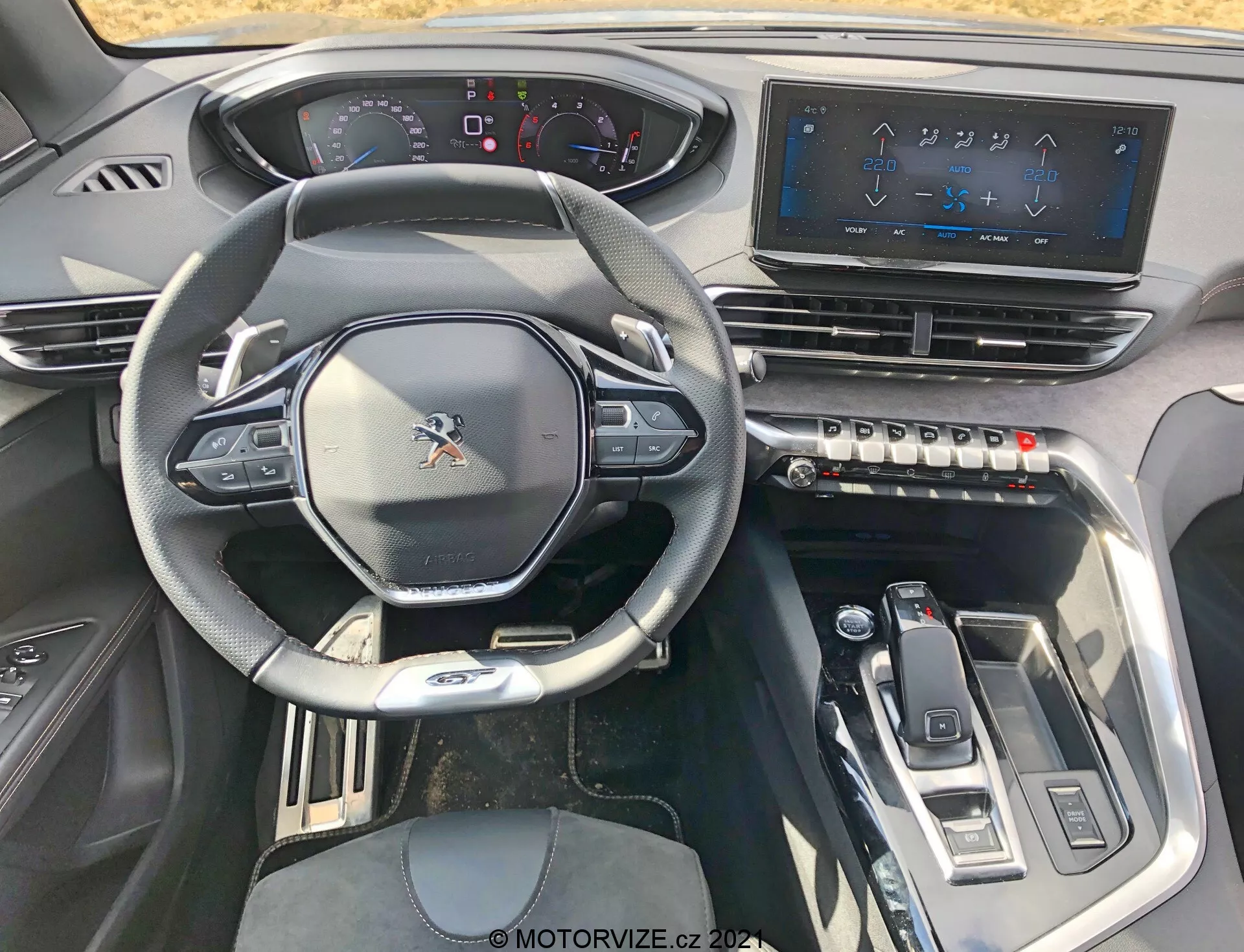 Vista interna di una Peugeot 5008 del 2021 dal sedile del conducente, che mostra il volante multifunzione sportivo con comandi montati, un cruscotto parzialmente digitale, un ampio sistema di infotainment con touchscreen, pulsanti tattili per il controllo del clima, un selettore del cambio elegante e elementi di design moderni con accenti metallici per un'esperienza high-tech e di alta gamma.