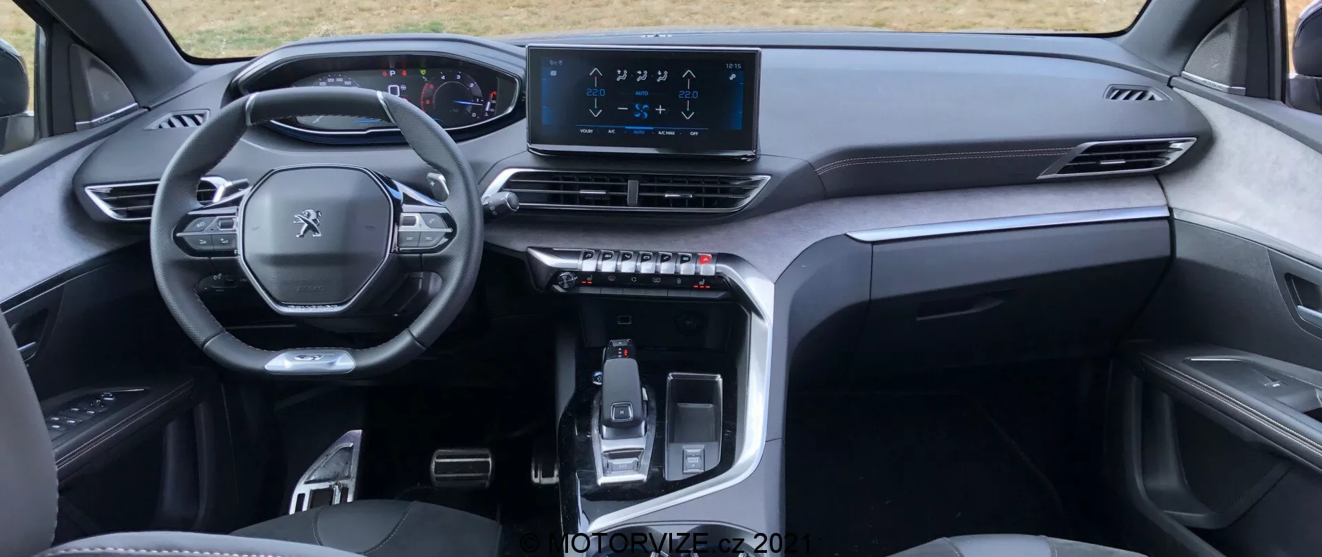 Vista interna di una Peugeot 5008 2021 dalla prospettiva del sedile posteriore, evidenziando un volante multifunzione, un cruscotto digitale, una plancia simmetrica con bocchette d'aria, uno schermo touchscreen centrale per l'infotainment e una console centrale elegante con leva del cambio controllata elettronicamente e pulsanti tattili. Il design minimalista e raffinato utilizza materiali di alta qualità, suggerendo un veicolo moderno e di categoria premium.
