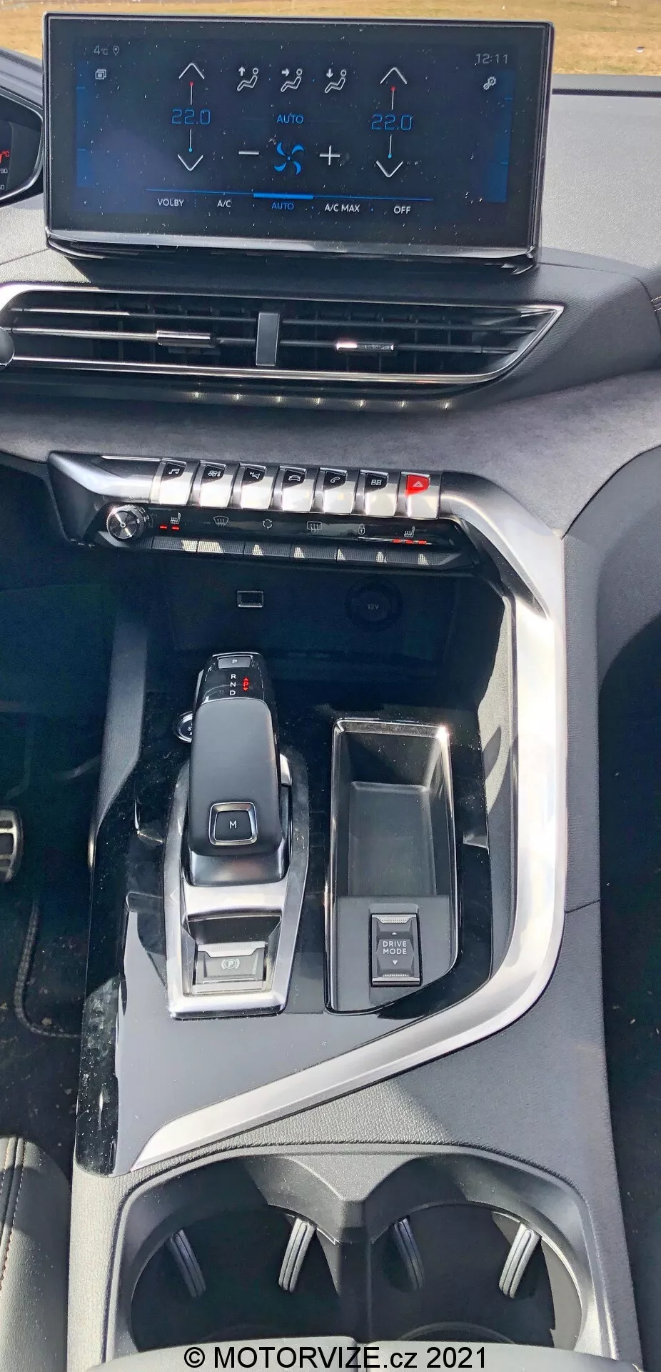 Vista dell'interno della Peugeot 5008 del 2021 che mostra la console centrale da una prospettiva dall'alto. L'immagine mette in evidenza un display infotainment touch screen flottante che mostra i comandi dell'aria condizionata sopra ai pulsanti e manopole fisici per il controllo del clima. Più in basso è visibile una leva del cambio elettronica elegante e i controlli circostanti per la selezione delle modalità di guida e una manopola di interfaccia multifunzionale. Il design suggerisce un veicolo moderno e premium con caratteristiche tecnologiche avanzate ed ergonomiche, inclusi portabicchieri e spazi di stoccaggio.