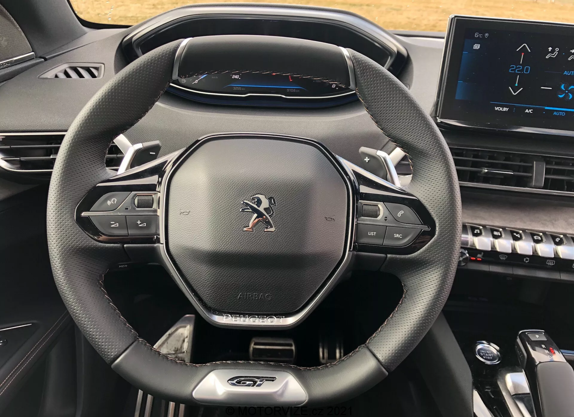 Vista ravvicinata dell'abitacolo del conducente all'interno di una Peugeot 5008 del 2021, evidenziando il moderno volante a tre razze con texture simile alla pelle, pulsanti di controllo multimediale e del cruise control, emblema Peugeot, etichetta 'AIRBAG', quadro strumenti completamente digitale che mostra un tachimetro con una velocità massima di 220 km/h e parte della console centrale con tasti stile pianoforte e un touchscreen elettronico, che mostra un design contemporaneo e tecnologicamente avanzato.