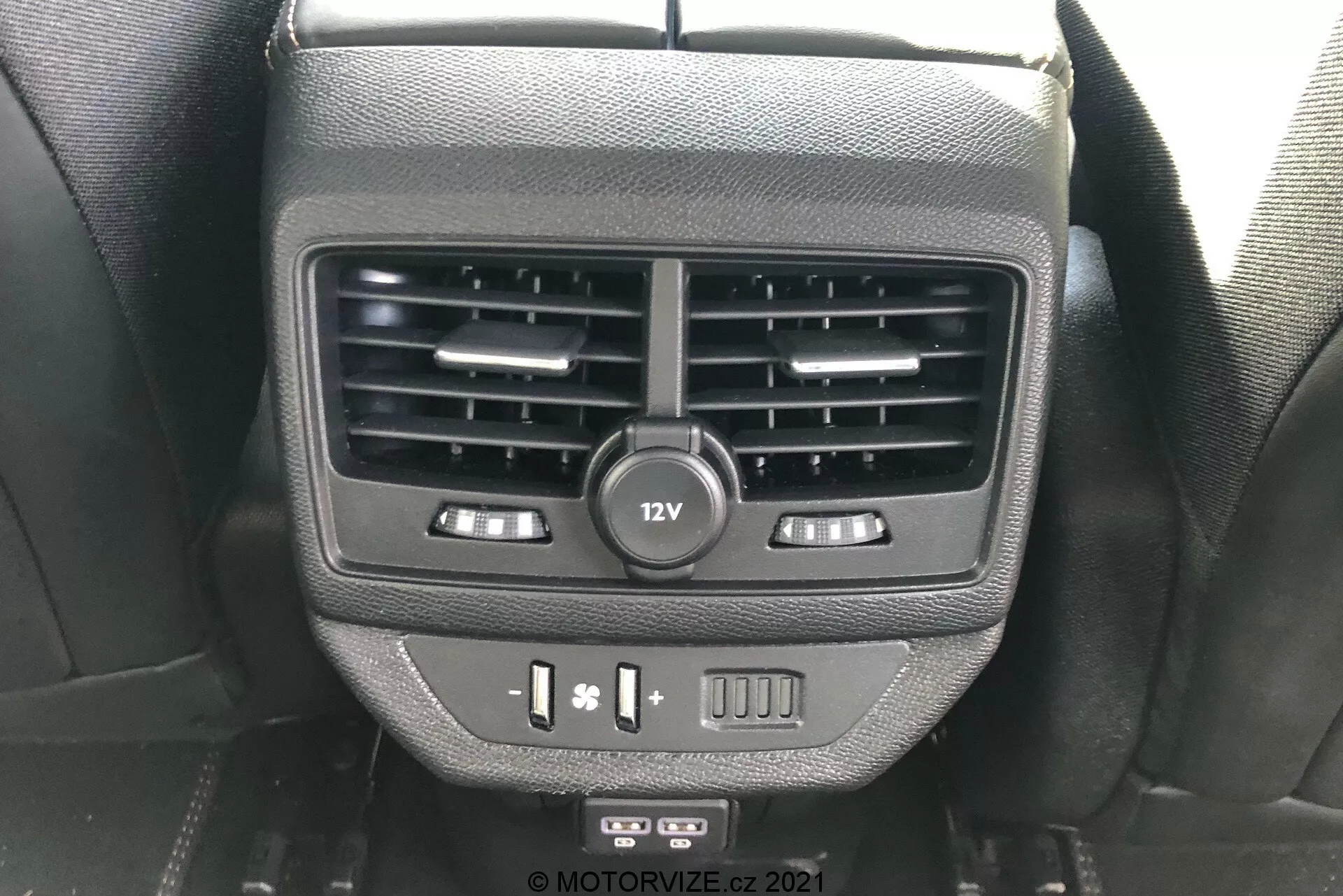 Vista ravvicinata delle caratteristiche per i passeggeri posteriori sul retro della console centrale della Peugeot 5008 2021. L'immagine mostra due bocchette d'aria, una presa di corrente da 12V, controlli del riscaldamento dei sedili, porte USB e un pulsante vuoto, catturata dalla prospettiva di un occupante del sedile posteriore.