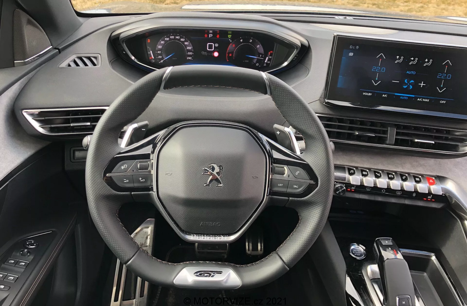 Vista interna della Peugeot 5008 2021 dal sedile del conducente, evidenziando il volante rivestito in pelle con fondo piatto con controlli integrati, un cruscotto completamente digitale con indicatori illuminati, un grande sistema di infotainment con touchscreen sulla console centrale, comandi del climatizzatore eleganti, selettore del cambio elettronico per trasmissione automatica e design di materiali scuri di alta qualità che trasmette un'estetica moderna, high-tech e premium.