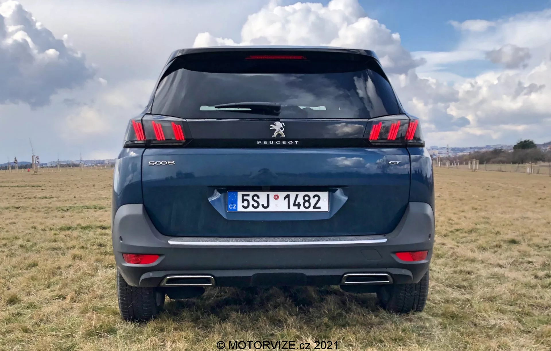 Vista posteriore di un moderno SUV Peugeot 5008, che evidenzia i raffinati fanali posteriori orizzontali con segmenti LED a forma di artiglio, il logo Peugeot e l'iscrizione del modello '5008'. Colore blu scuro o grigio, parcheggiato su un terreno esterno con un cielo parzialmente nuvoloso. Gli elementi visibili includono il parabrezza posteriore, il tergicristallo posteriore, la luce stop montata in alto, la maniglia del portellone e la targa della Repubblica Ceca con 'CZ'. Striscia riflettente sopra il paraurti con sensori di parcheggio e ritagli di terminali di scarico evidenti. Scatto centrato con l'auto che riempie la maggior parte dell'inquadratura.