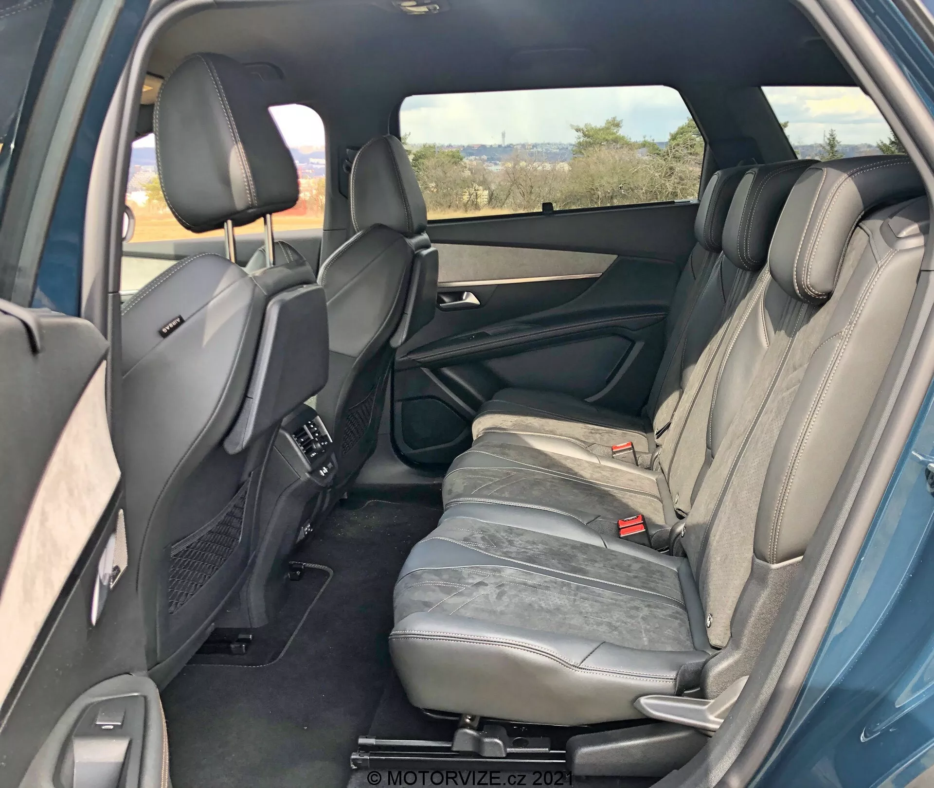 Interni di una Peugeot 5008 2021 dalla prospettiva posteriore destra che mostra il compartimento posteriore per i passeggeri. La porta posteriore destra è aperta rivelando le caratteristiche del pannello della porta come maniglia, scomparti di stoccaggio e altoparlante. Sono visibili tre sedili posteriori rivestiti in pelle grigia con poggiatesta. Il bracciolo del sedile centrale è in posizione verticale. Sono anche visibili le cinture di sicurezza posteriori e i sedili anteriori con poggiatesta regolabili e consolle centrale. Bocchette di controllo del clima posteriori sulla consolle centrale, finestrino destro oscurato, scorci della porta posteriore sinistra che indicano simmetria. In generale, interni ben curati senza caratteristiche tecnologiche visibili.