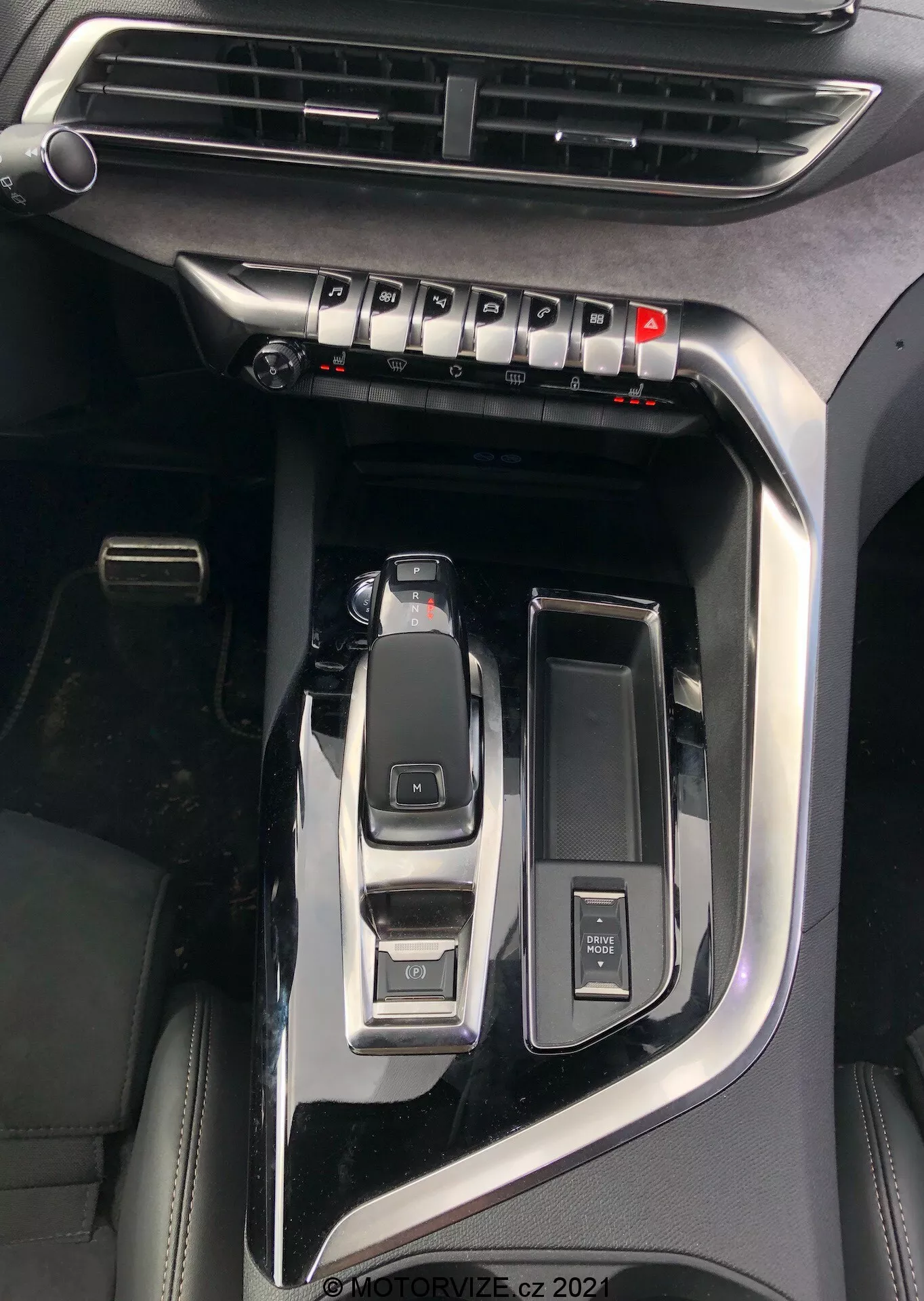 Inquadratura ravvicinata della console centrale della Peugeot 5008 2021 con evidenza dei principali elementi come la leva del cambio automatico in posizione di parcheggio, il selettore delle modalità di guida per modalità eco, sport e comfort, un pulsante di avviamento/arresto del motore per l'accensione senza chiave, una manopola di controllo multimediale, un porta bicchieri aperto, una bocchetta dell'aria visibile per il controllo del clima e una finitura in nero pianoforte con accenti metallici che indicano un modello premium, filigrana 'MOTORVIZE.cz 2022'.