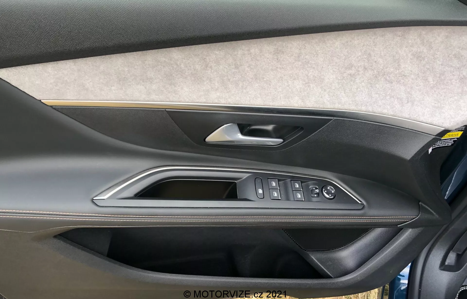 Vista ravvicinata del pannello della portiera interna del Peugeot 5008 2021 con una sezione rivestita in feltro o tessuto, un design in plastica modellata bicolore con una parte superiore più scura e una parte inferiore più chiara, maniglia della porta in materiale di alta qualità metallico o cromato, finiture lucidate e comandi ergonomici per i finestrini elettrici, chiusura delle porte e specchietti laterali. È visibile anche una tasca portaoggetti nella parte inferiore del pannello della porta.