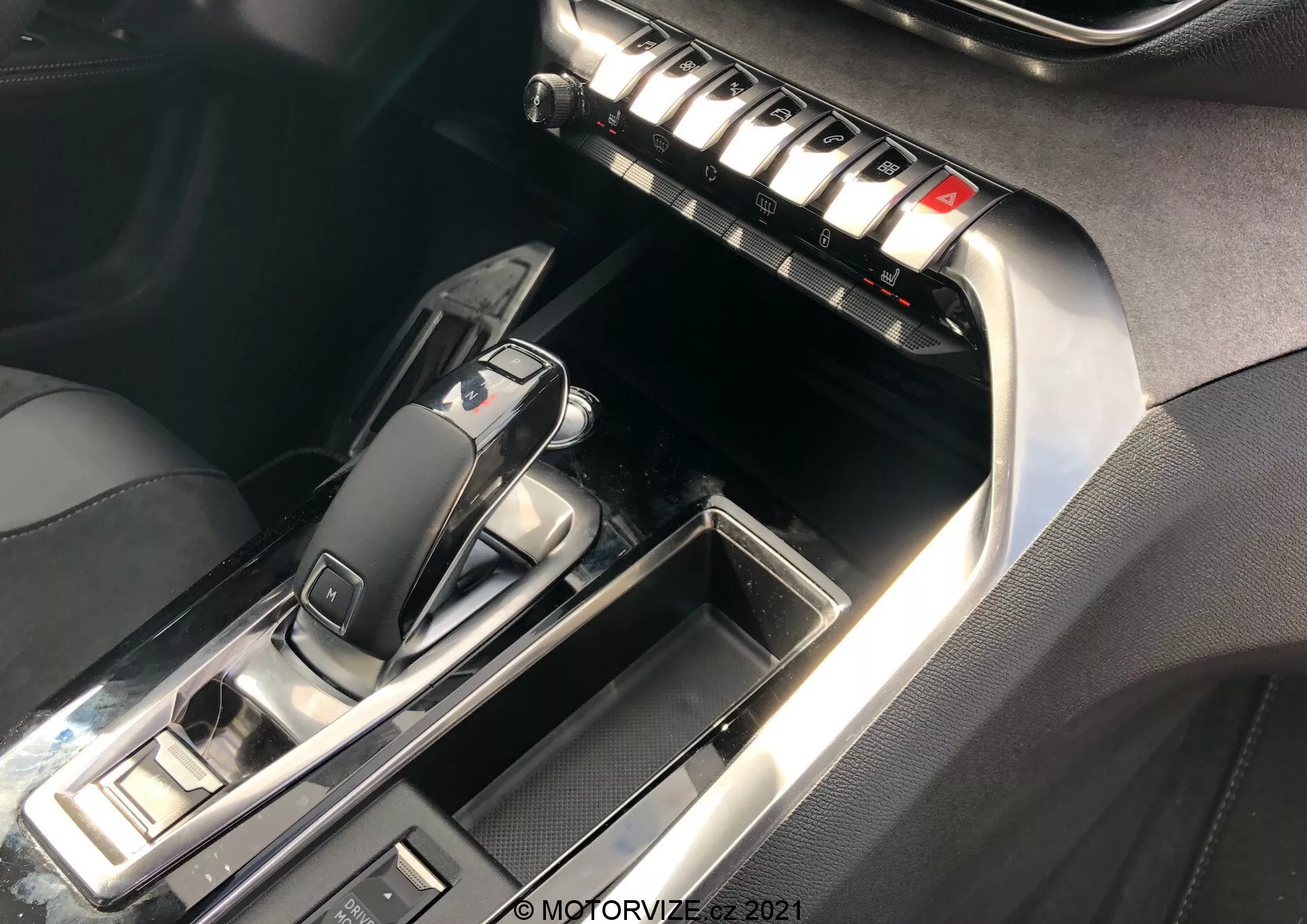 Vista ravvicinata della consolle centrale e dell'interno anteriore della Peugeot 5008 2021, focalizzandosi sull'area del cambio automatico e sul pannello centrale. La leva del cambio, in posizione 'P', presenta un design elegante con plastica nera lucida e finiture metalliche. I pulsanti circostanti suggeriscono modalità di guida e controlli del freno di stazionamento. Il pannello centrale include una fila di pulsanti metallici probabilmente per il controllo del clima, gli sbrinatori e le impostazioni dei sedili. Cuciture a contrasto su pelle o similpelle e accenti cromati aumentano la sensazione di lusso. L'illuminazione enfatizza le superfici riflettenti e i dettagli.