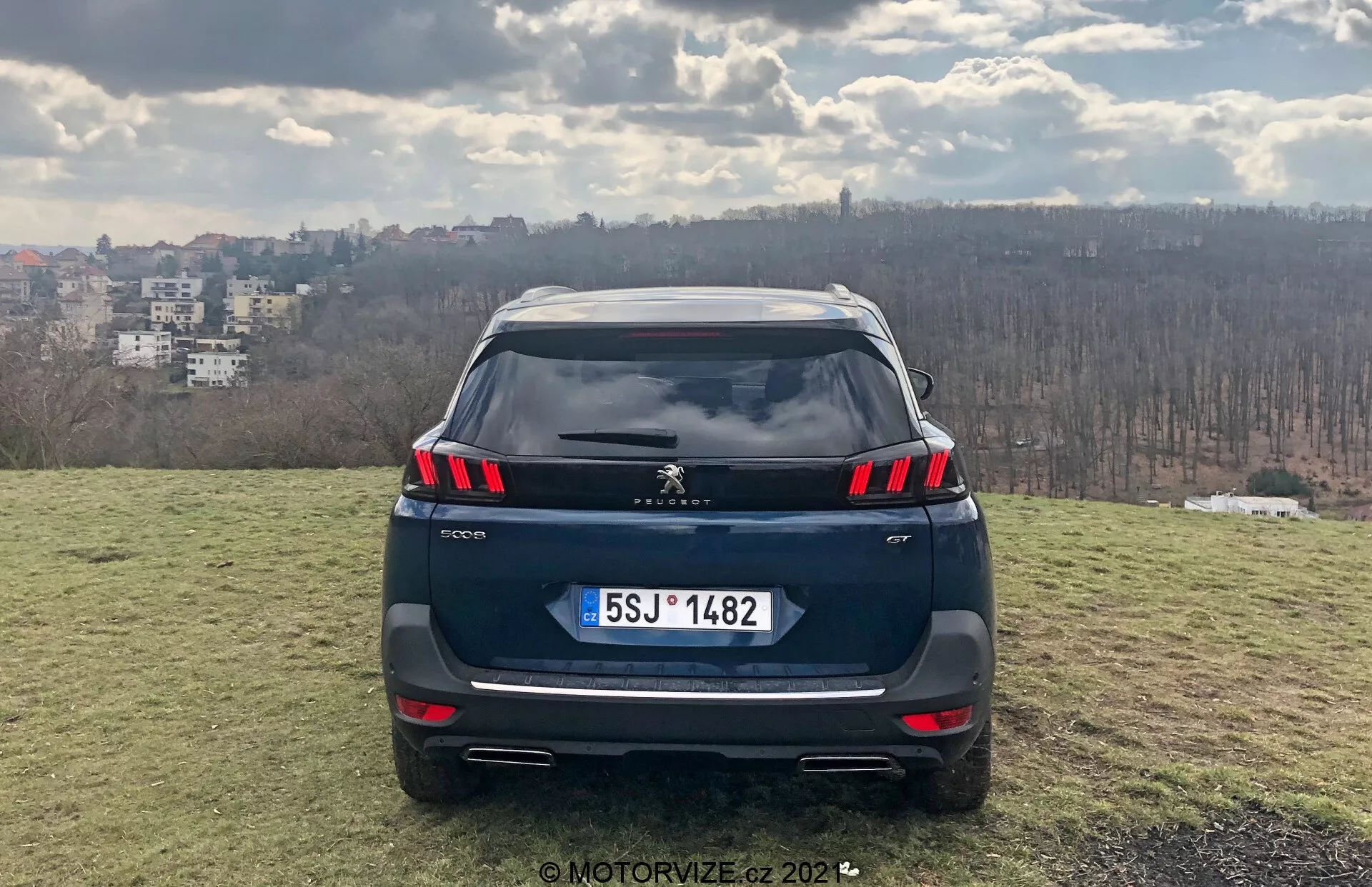Vista posteriore frontale di un SUV Peugeot 5008 2021 su un'area erbosa elevata con uno skyline cittadino sullo sfondo, mostrando gruppi ottici posteriori a LED, sensori di parcheggio integrati nel paraurti, possibile retrocamera, targa in stile europeo, doppi terminali di scarico, spoiler sul tetto e antenna a pinna di squalo; ambientazione diurna che suggerisce parziale nuvolosità, fotografato da MotorVize.cz nel 2022.