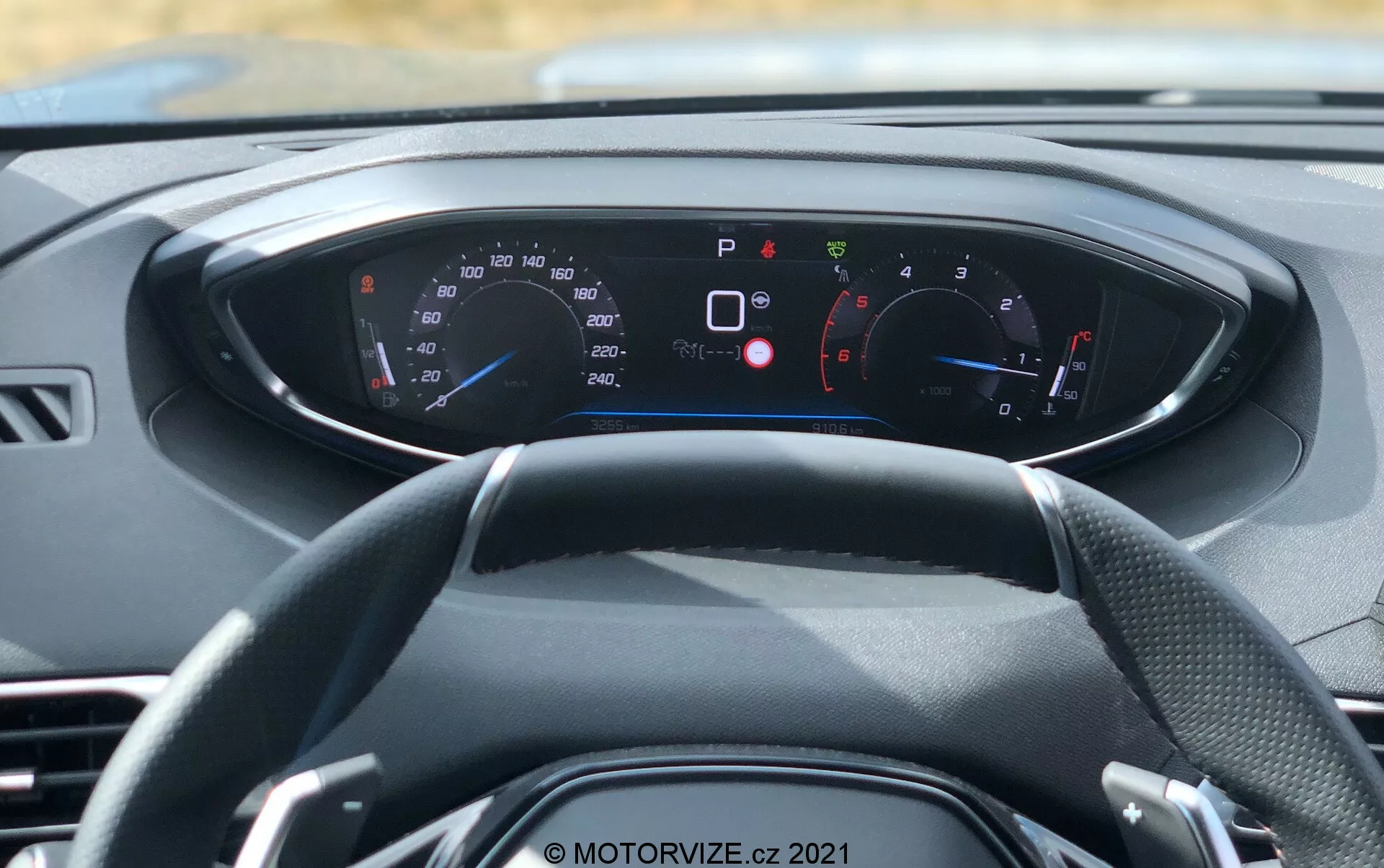 Vista interna di un veicolo moderno da una prospettiva in prima persona, con attenzione al volante e al cruscotto digitale. Il volante a tre razze è centrato, con vari indicatori e display illuminati sul cruscotto dietro di esso. Il tachimetro e il contagiri sono visibili, con letture in km/h e mph, adatti per mercati internazionali. Il cruscotto mostra numerose spie di avviso e informazioni, come freno di stazionamento inserito, porte aperte, promemoria cintura di sicurezza e pressione bassa degli pneumatici, indicando caratteristiche avanzate di diagnostica e sicurezza a bordo. Lo schema di colori del cruscotto è blu e rosso su uno sfondo nero con testo bianco. Questa immagine, scattata alla luce del giorno, enfatizza il design degli interni dell'auto, la strumentazione digitale e la tecnologia moderna.