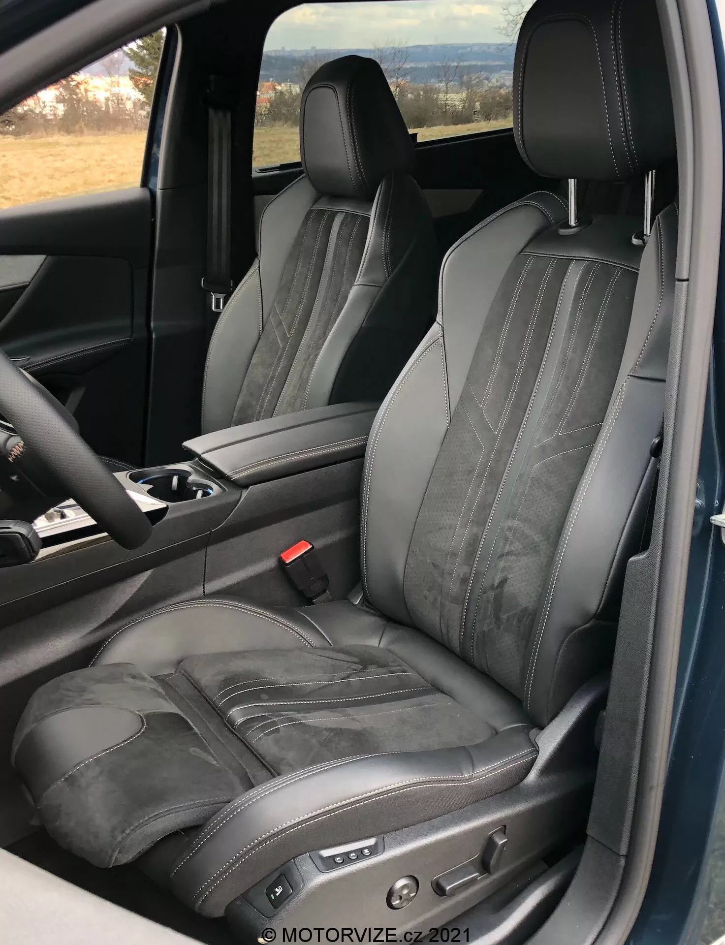 Vista dell'interno della Peugeot 5008 2021 incentrata sul sedile lato guida, che mostra un sedile regolabile in pelle nera con cuciture a contrasto e goffratura orizzontale, scattata da una prospettiva a porta aperta, evidenziando il design ergonomico e di lusso con comandi elettronici di regolazione del sedile e una parte della console centrale e del volante visibile.