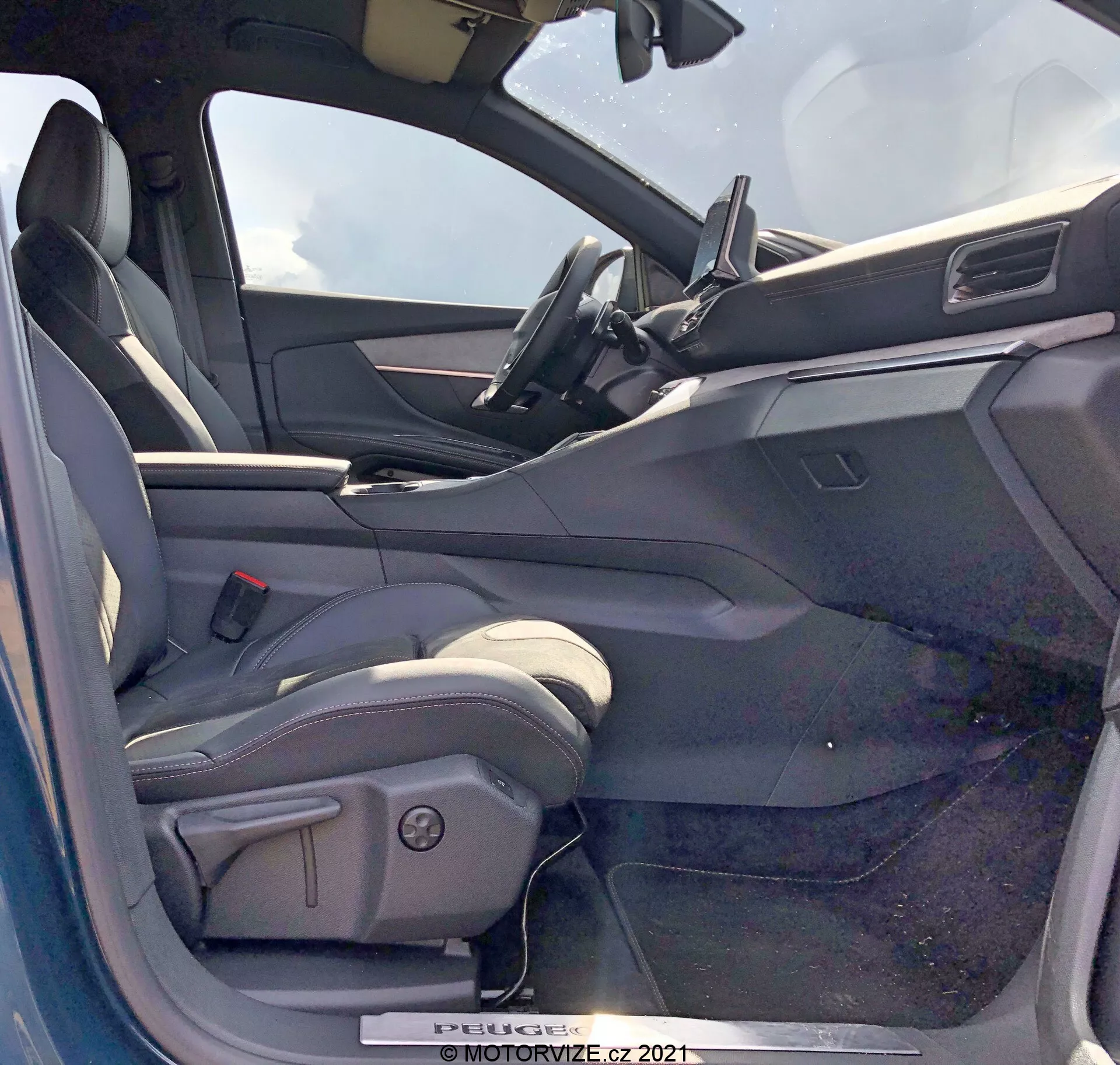Vista dell'interno di una Peugeot 5008 del 2021 dal lato del conducente con la portiera aperta. Il sedile del conducente è di tipo avvolgente, probabilmente rivestito in pelle o materiale simile alla pelle, con supporti laterali e comandi di regolazione elettrica. La portiera del conducente aperta mostra un pannello interno con comandi per i finestrini elettrici, probabilmente anche per gli specchietti elettrici e i meccanismi di chiusura della porta. Il cruscotto include uno schermo touch di infotainment posizionato centralmente con un design minimalista. La parte superiore del volante è visibile, suggerendo un volante rivestito in pelle con comandi integrati. L'interno ha uno schema di colori bicolore con superfici superiori più scure e superfici inferiori più chiare.